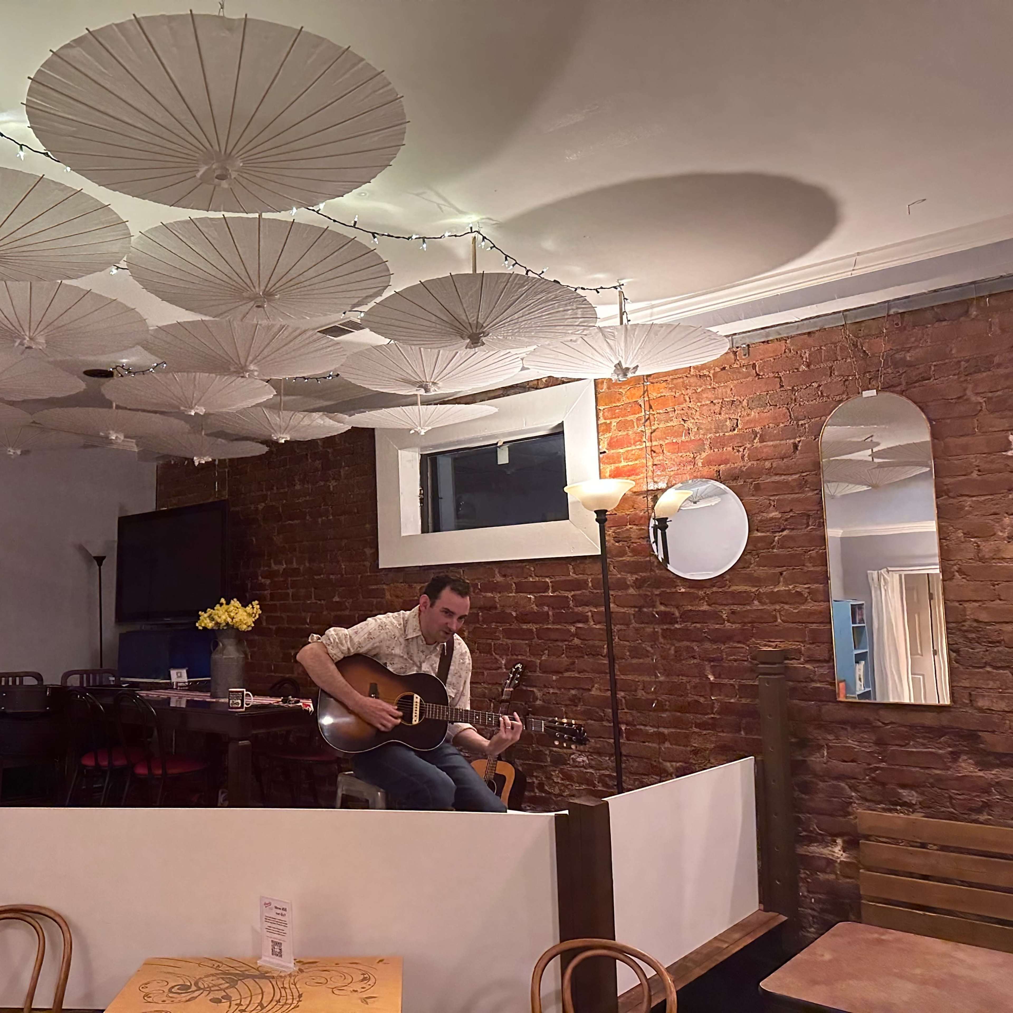 A musician plays guitar in a cozy café with a brick wall and decorative paper umbrellas hanging from the ceiling.