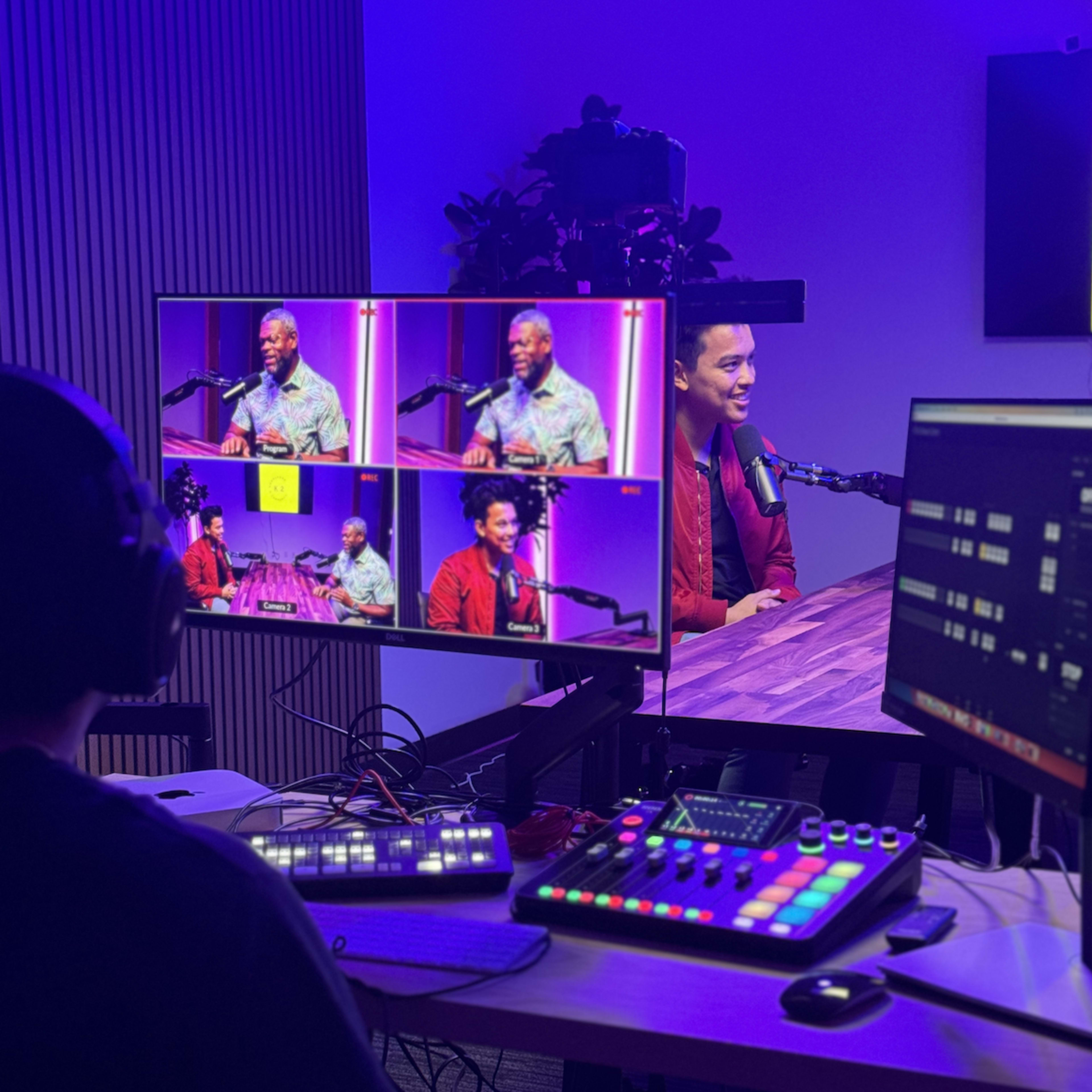 A person seated at a desk monitors multiple screens displaying a podcast recording with two speakers in front of microphones.