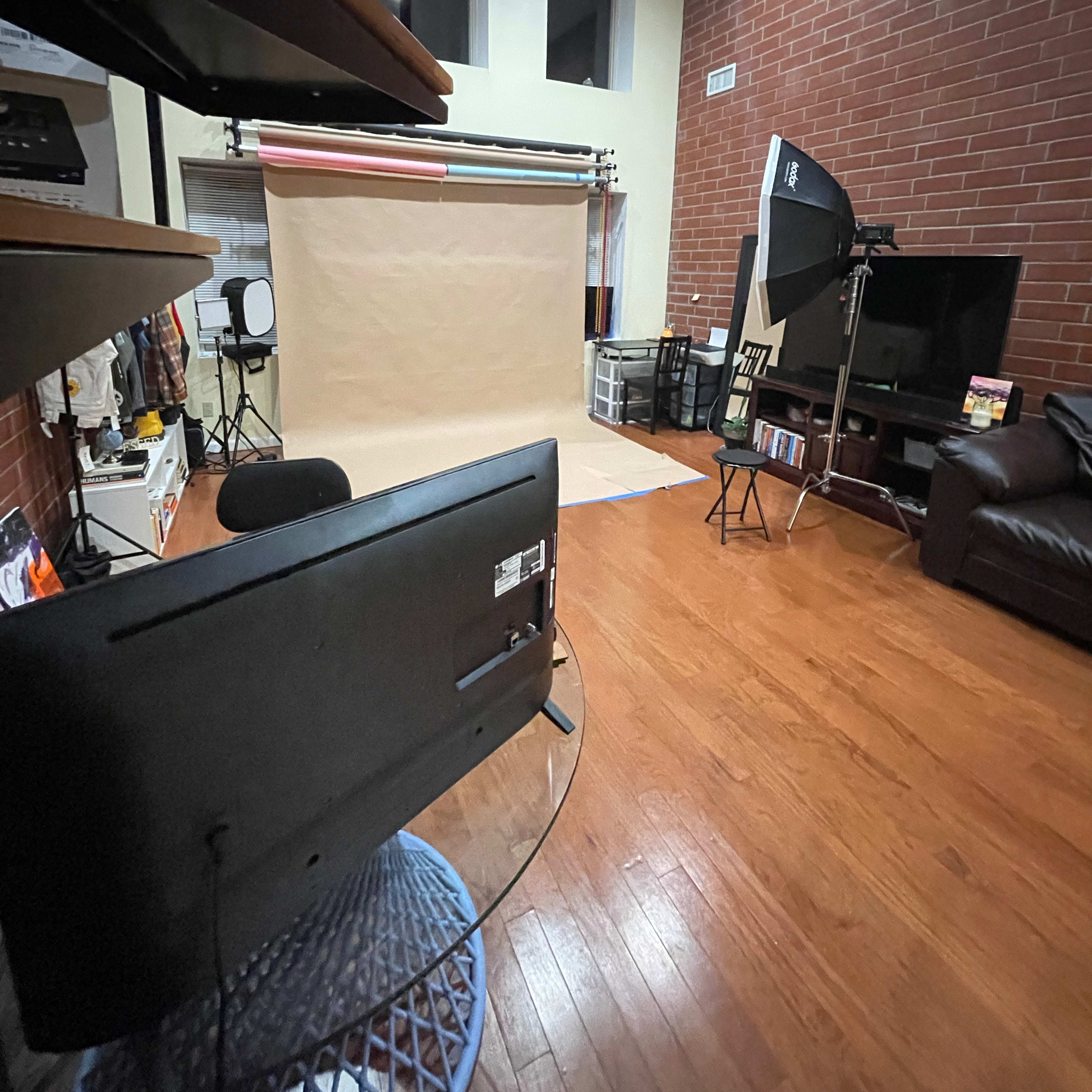 An interior studio setting with a seamless backdrop, lighting equipment, and a living area featuring a television and couch.
