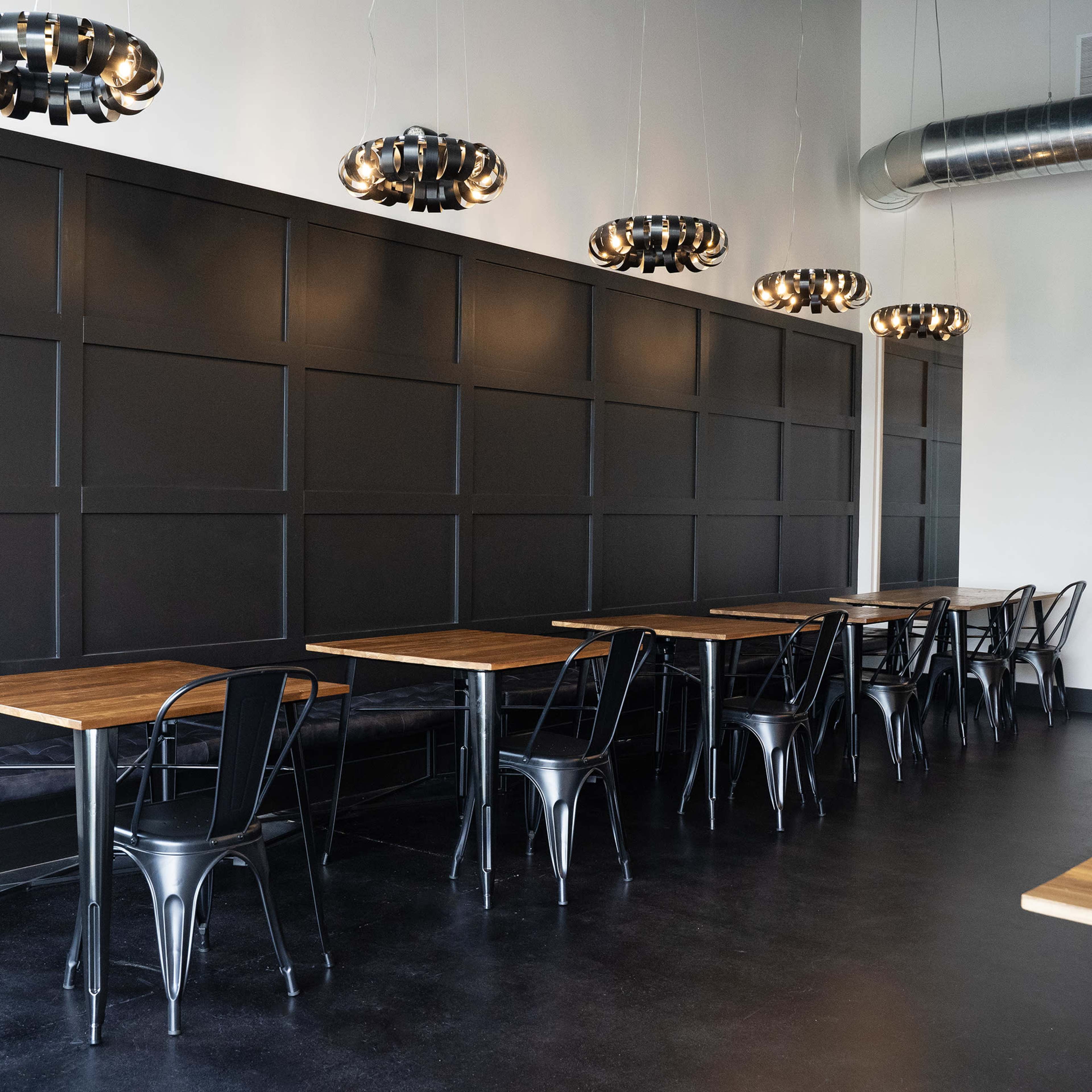 A modern, minimalist restaurant interior with black walls, wooden tables, and hanging light fixtures.
