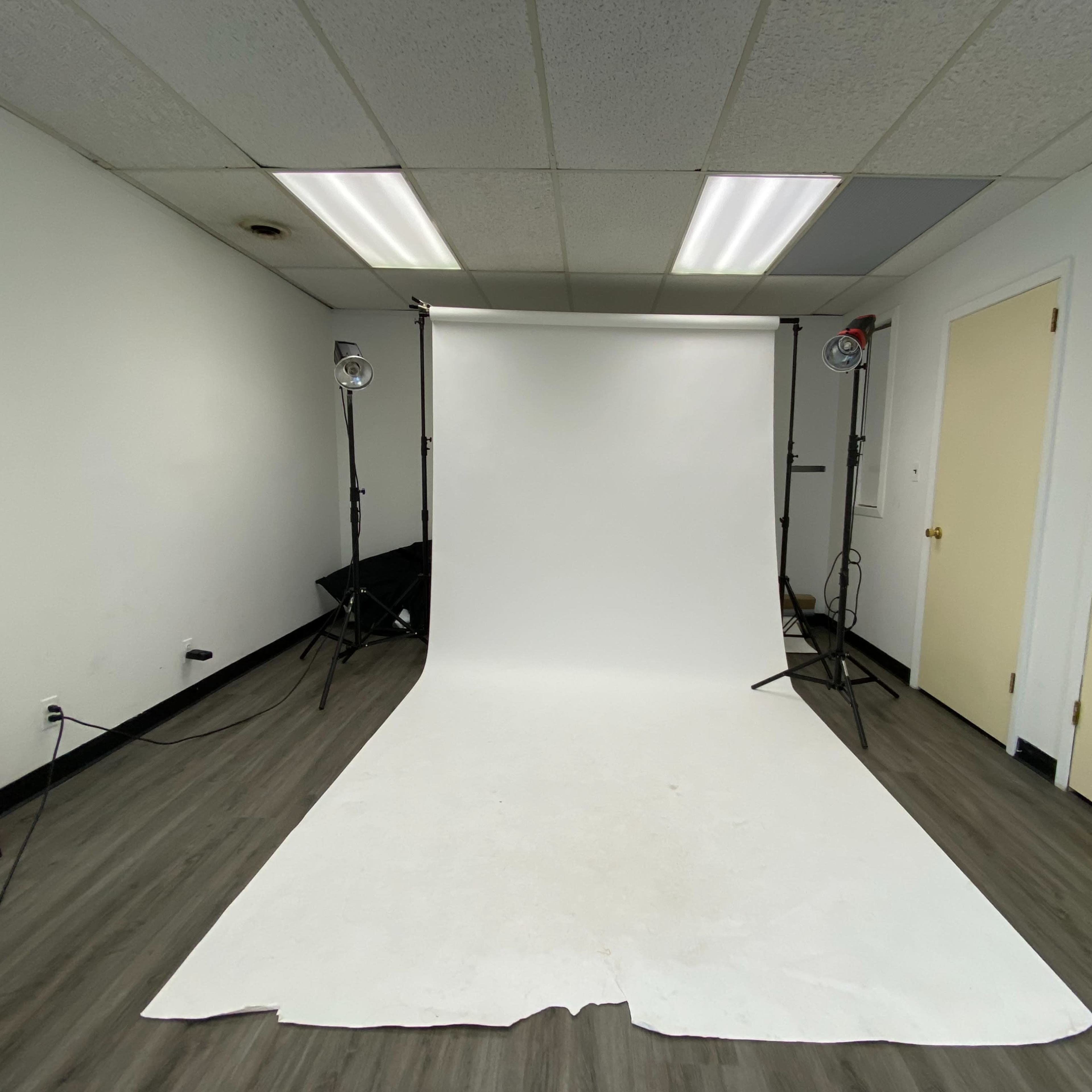 A photo studio with a white backdrop, two lights on stands, and a fan in the corner.