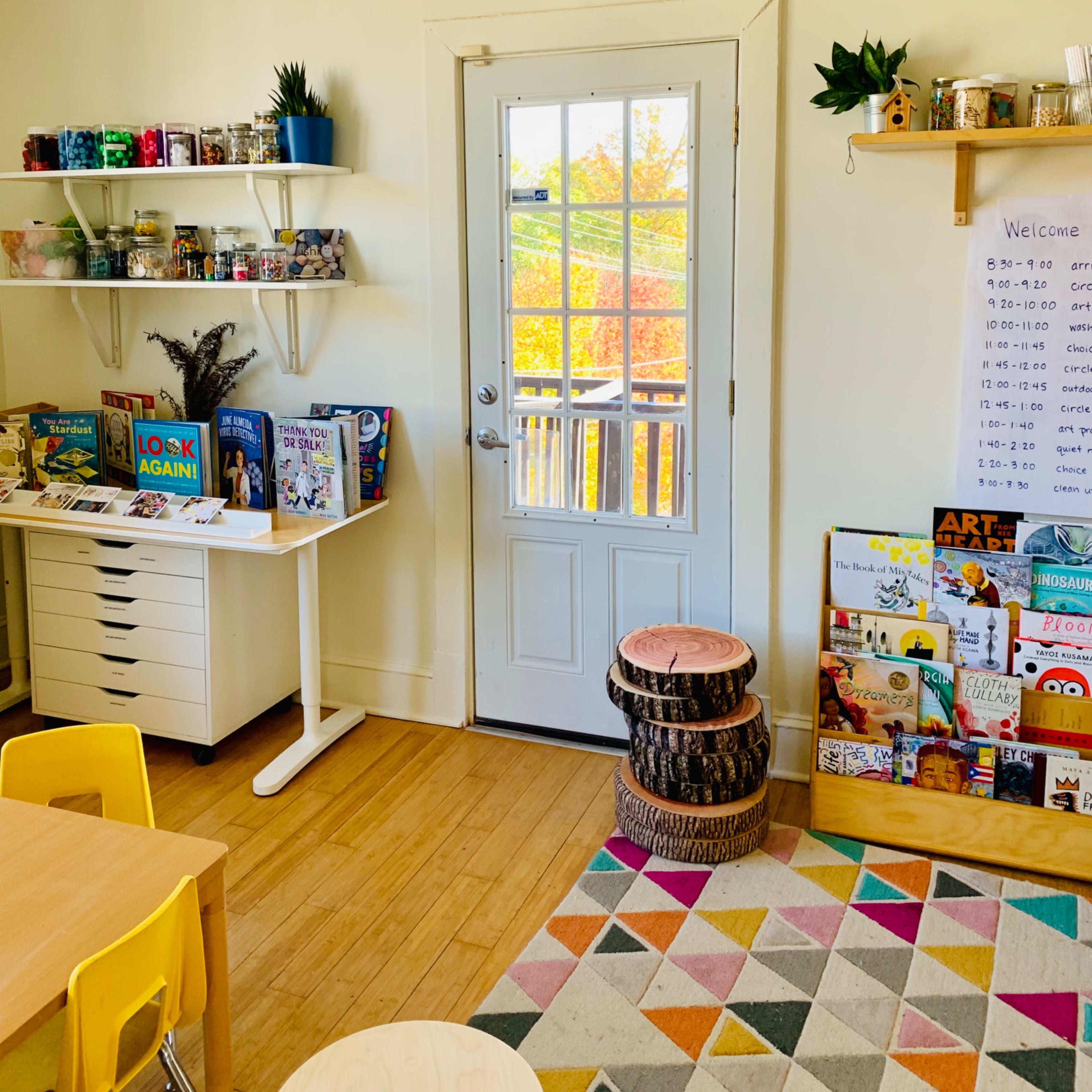A brightly lit room features a white door leading to an outdoor view, bookshelves stocked with colorful books and materials, and a patterned rug under a table and chairs.