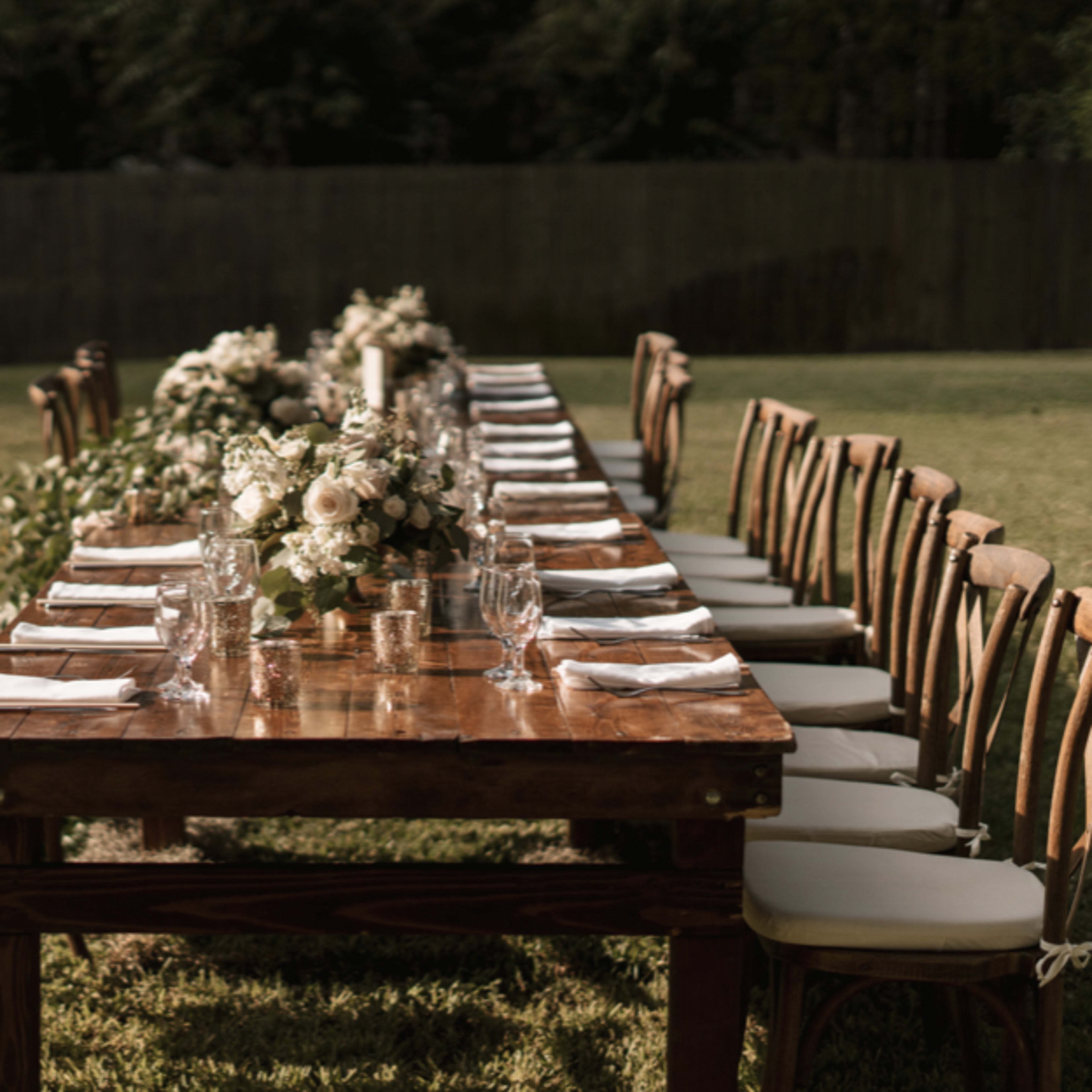 A long wooden dining table is arranged outdoors with neatly placed plates, glasses, and floral centerpieces.
