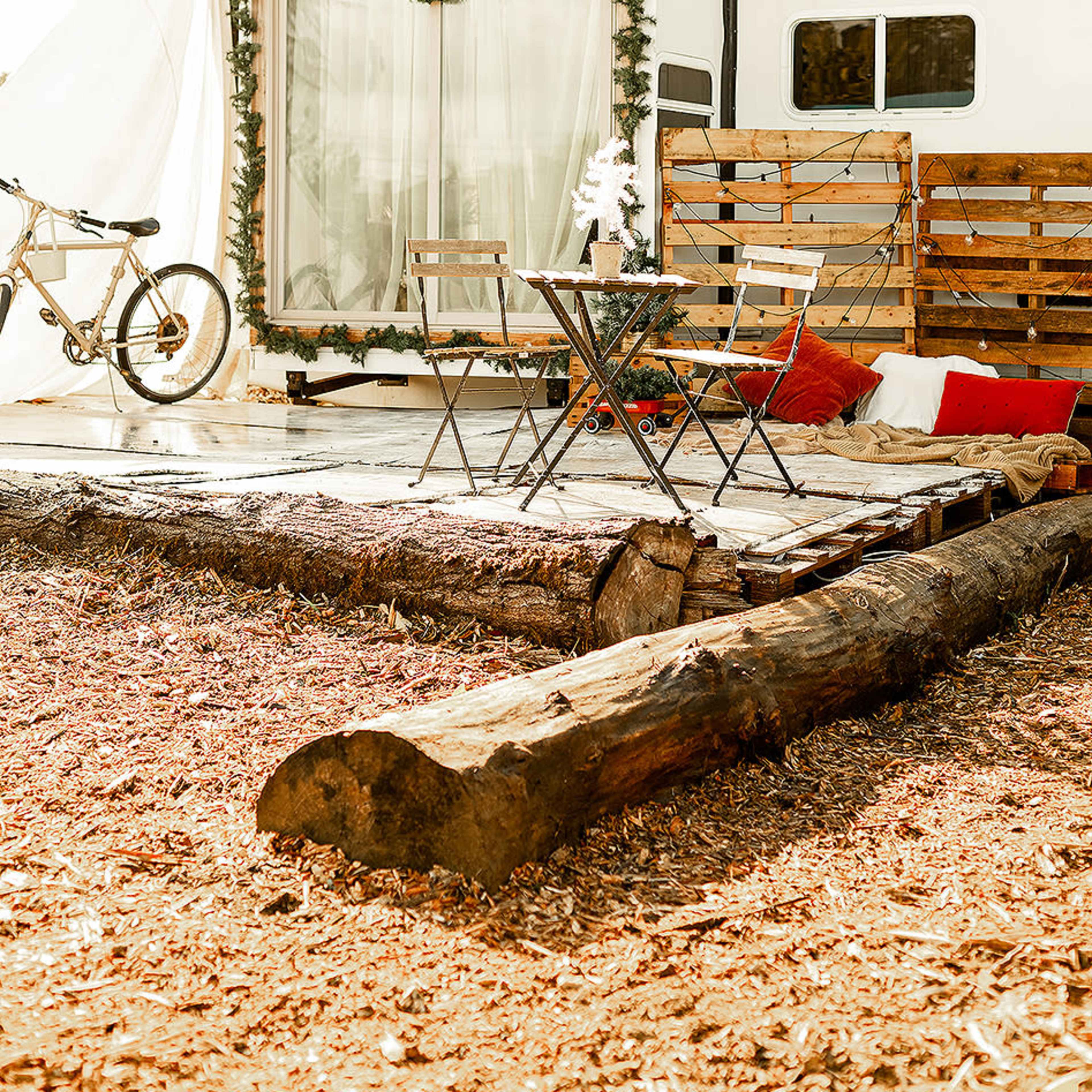 A rustic outdoor seating area with a small table and chairs, set beside a camper adorned with festive decorations and surrounded by wooden elements.