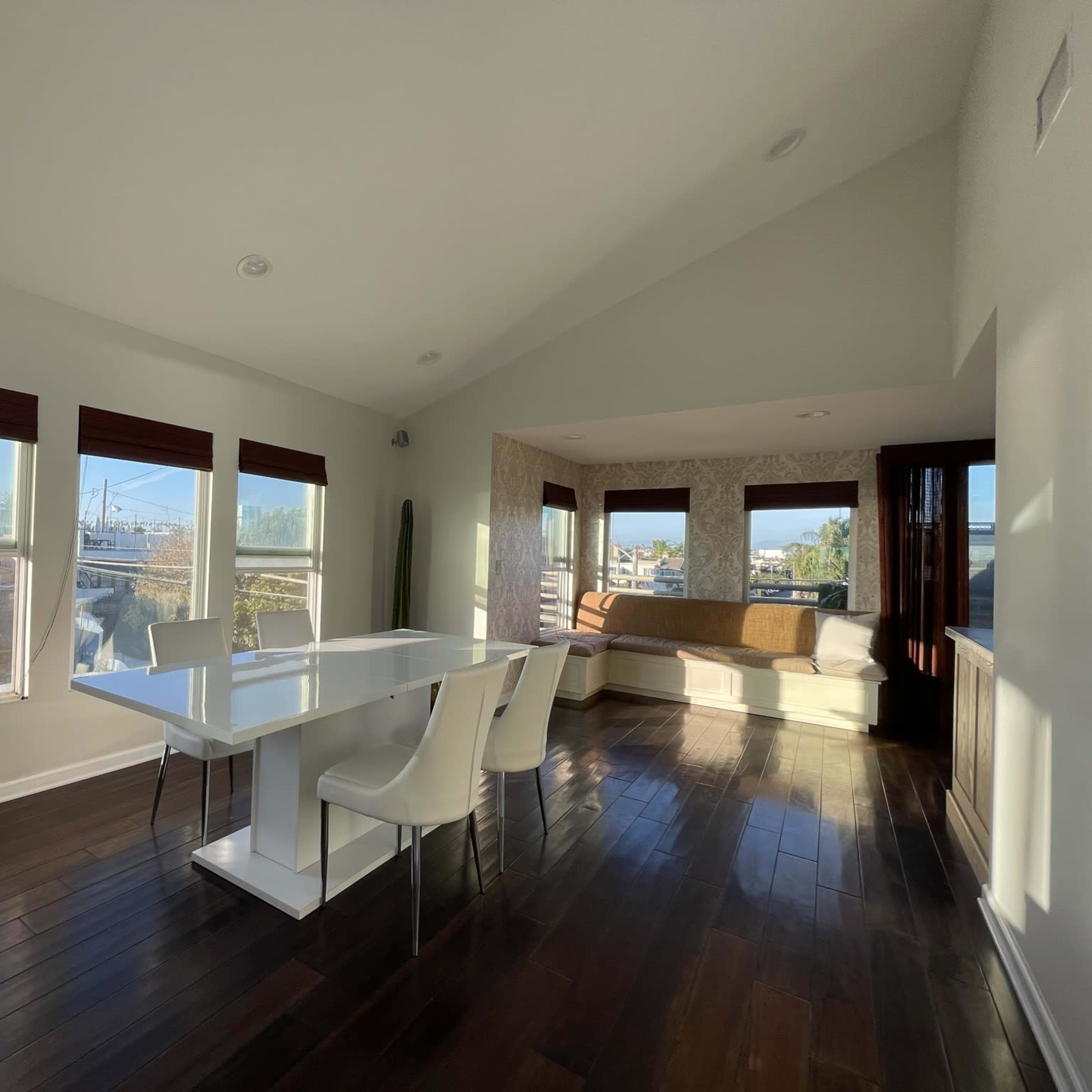 A modern dining area features a white table with four chairs, large windows allowing natural light, and a cozy seating nook in the corner.