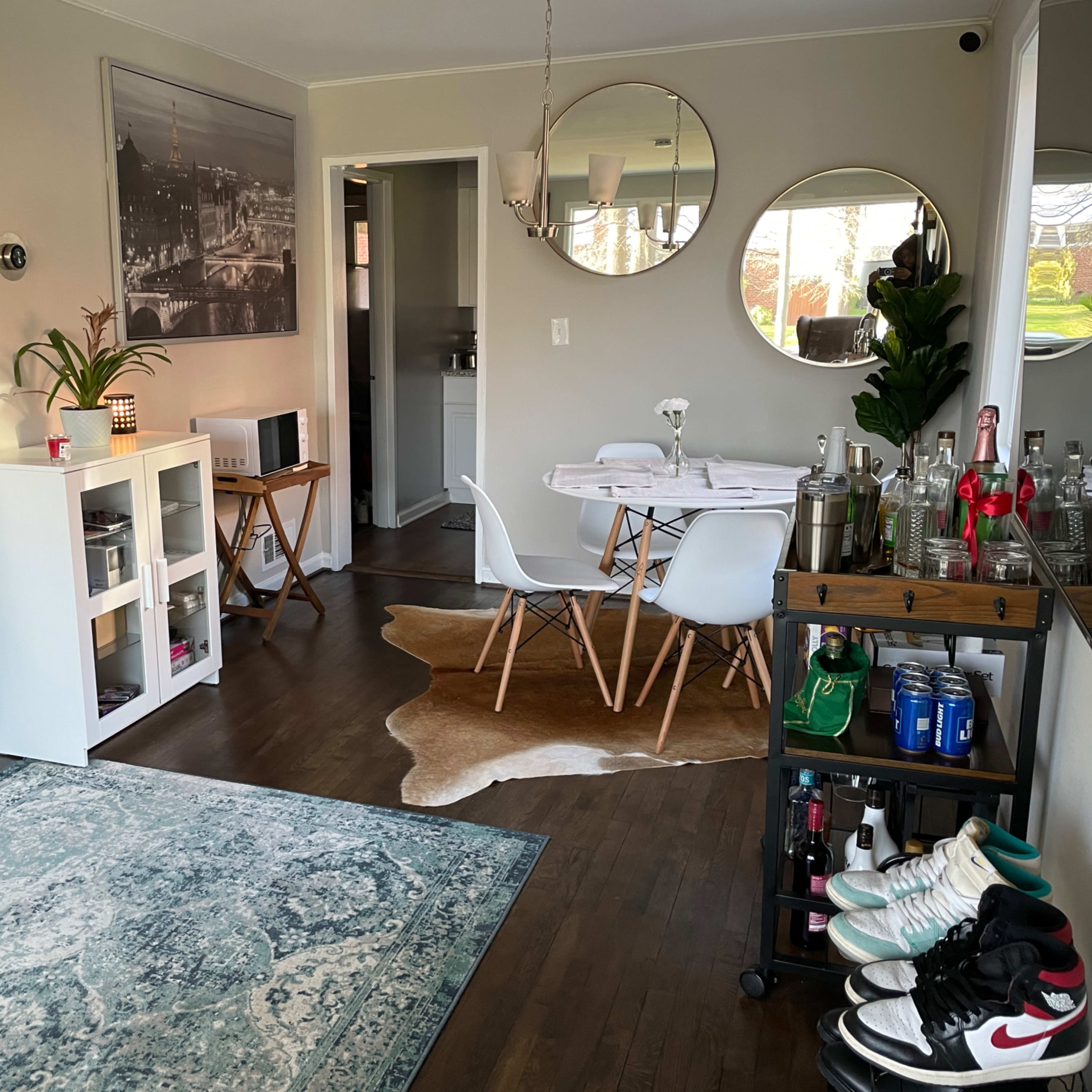 A modern living area features a green couch, a round dining table with white chairs, a bar cart, and decorative elements on the walls and floor.