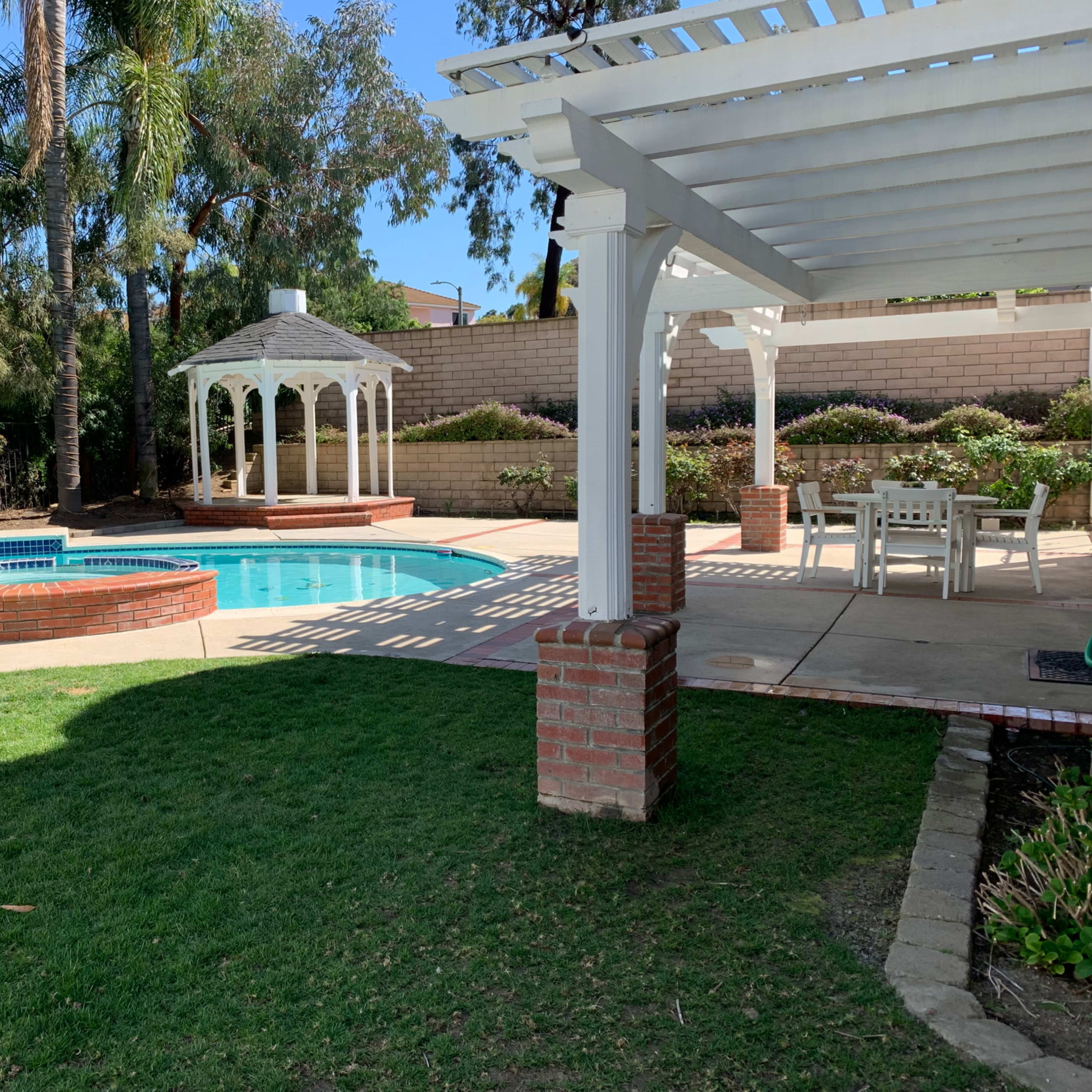 A backyard features a swimming pool with a spa, surrounded by palm trees, and includes a gazebo and a dining area under a pergola.