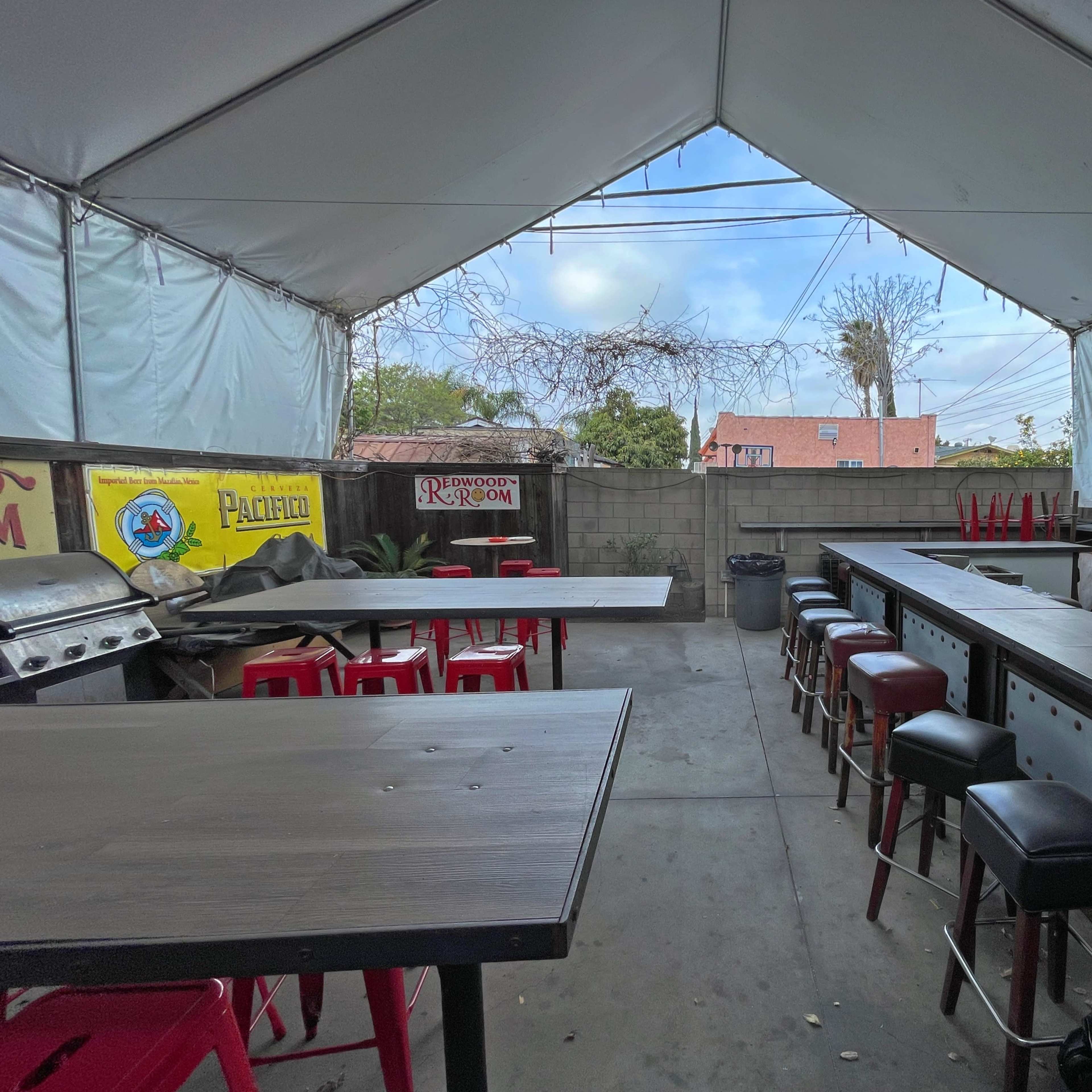 An outdoor covered space with several tables and bar stools, a grill, and empty walls, under a tent structure.