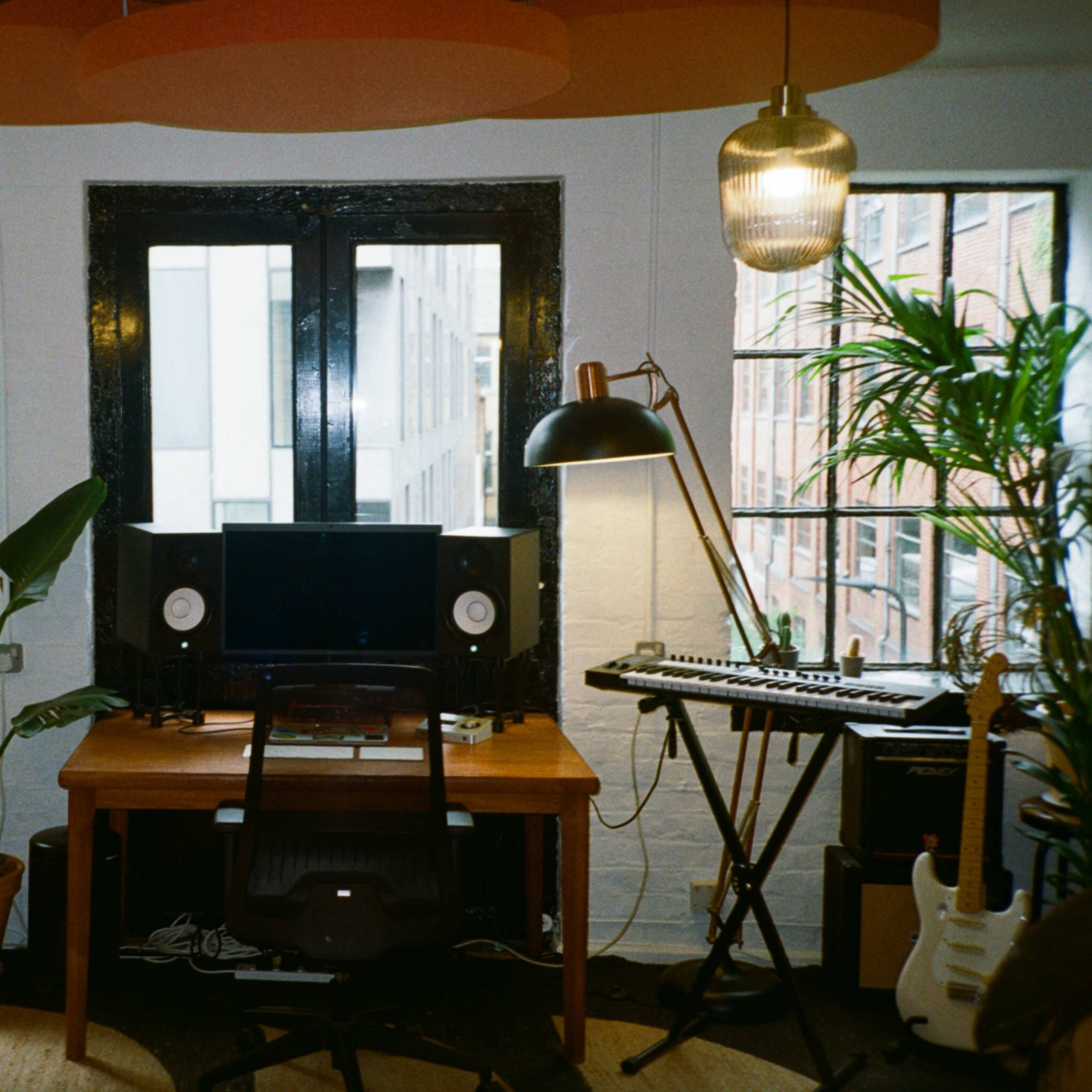 A modern workspace features a desk with a computer and speakers, surrounded by large windows, plants, a guitar, and a keyboard.