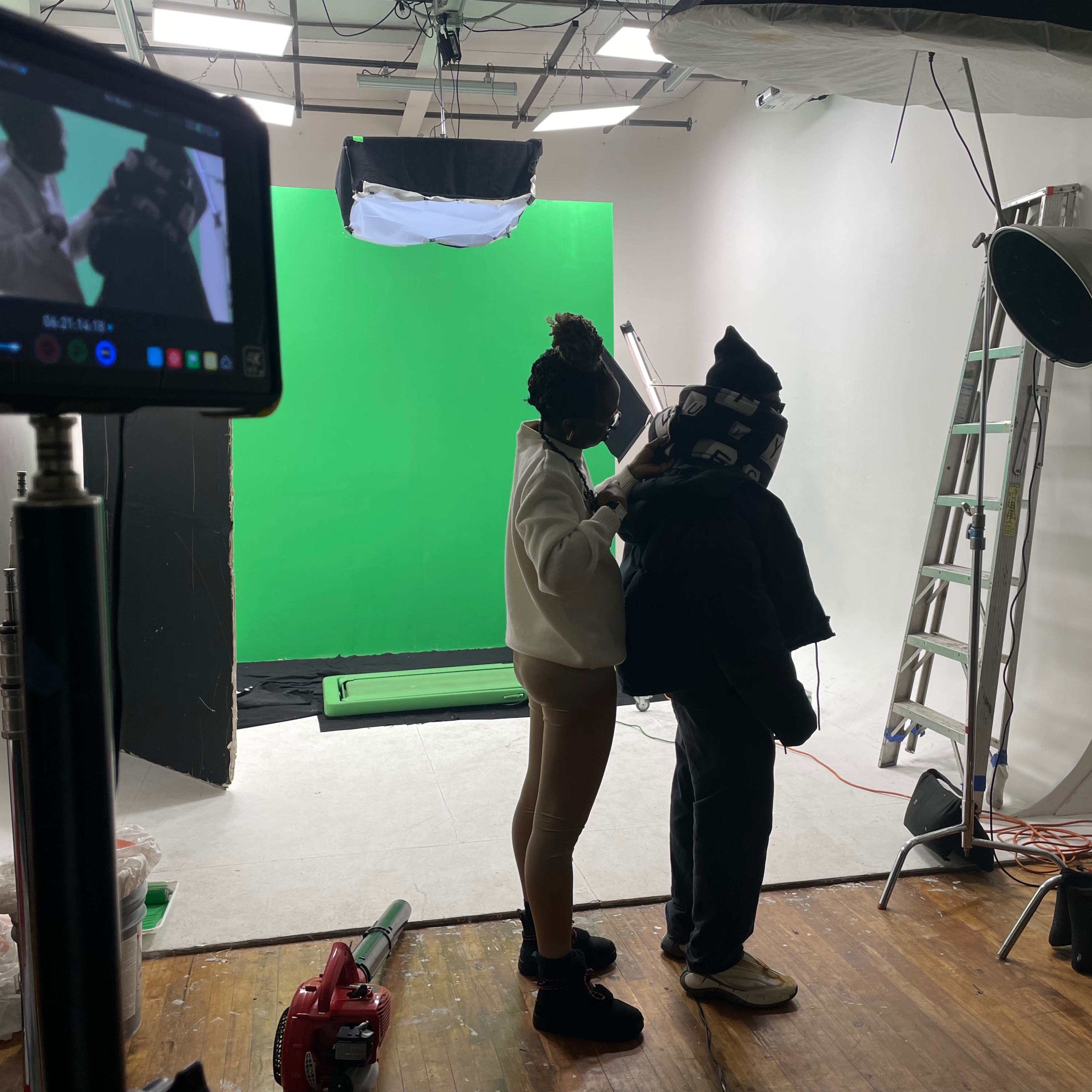 A person is adjusting clothing on another individual in a studio setting with a green screen and various lighting equipment.