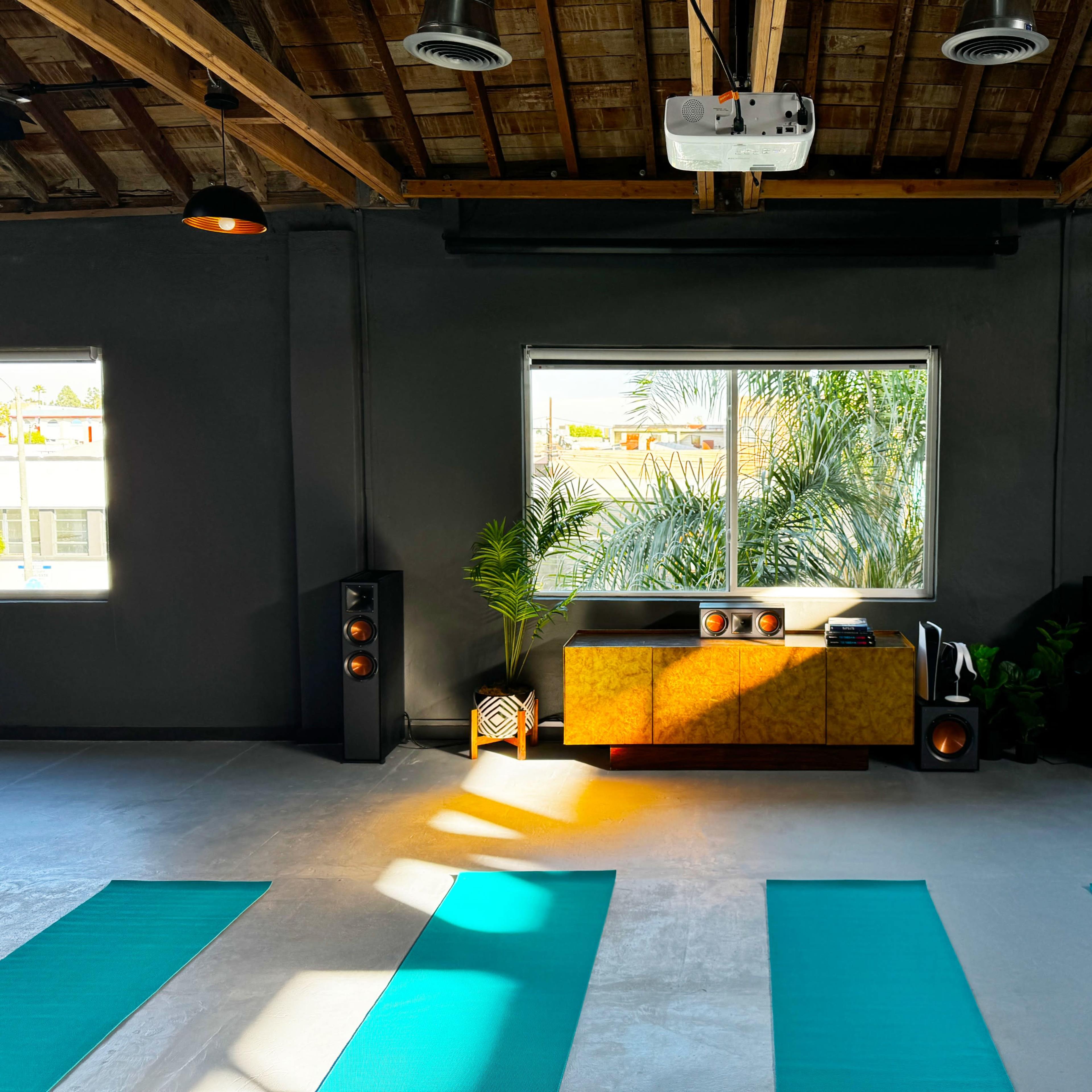 A spacious yoga studio with teal mats arranged on a concrete floor, a wooden cabinet under a window, and large speakers positioned on either side.