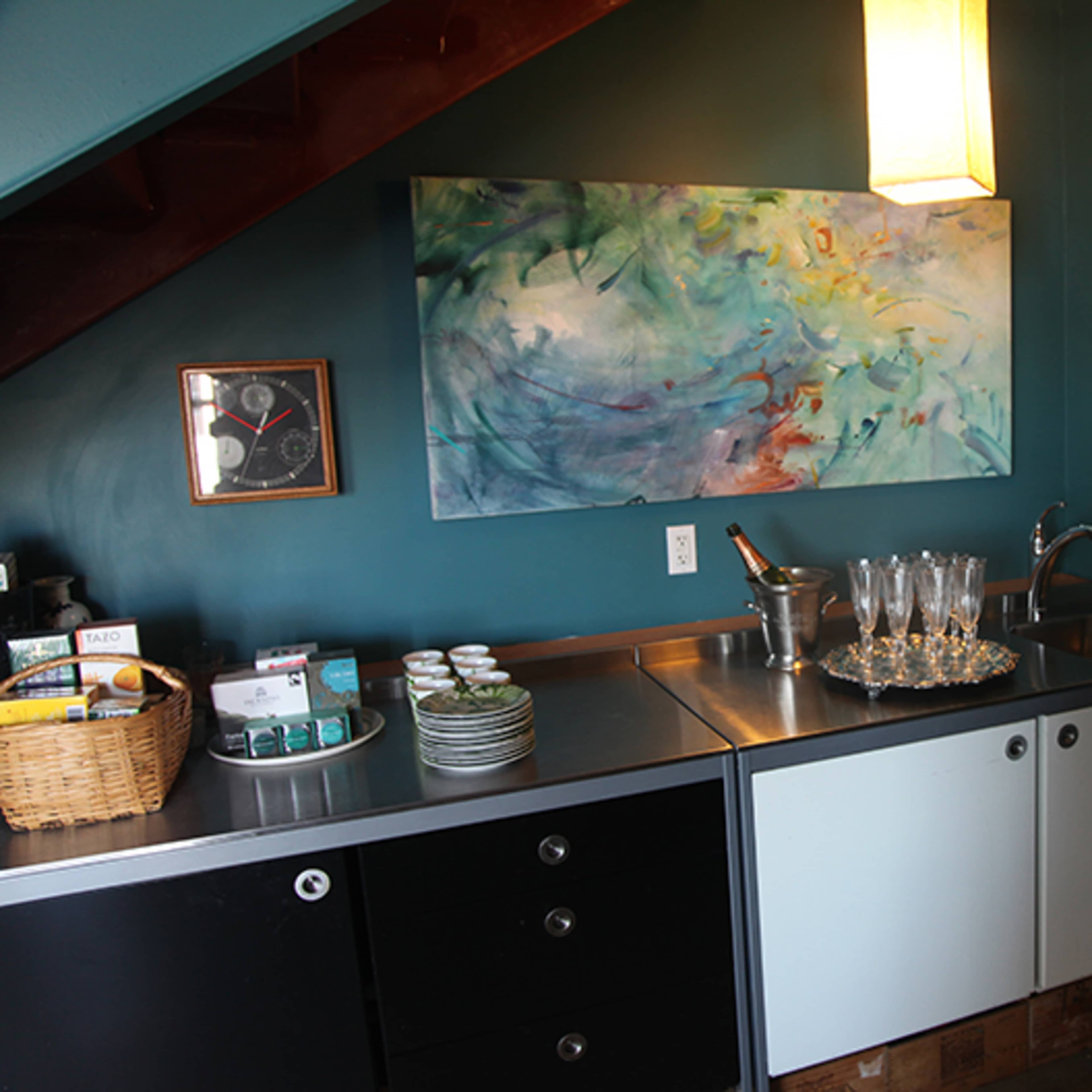 A compact kitchen features a silver sink counter with a basket of utensils, a selection of dishware, and an abstract painting on the wall.