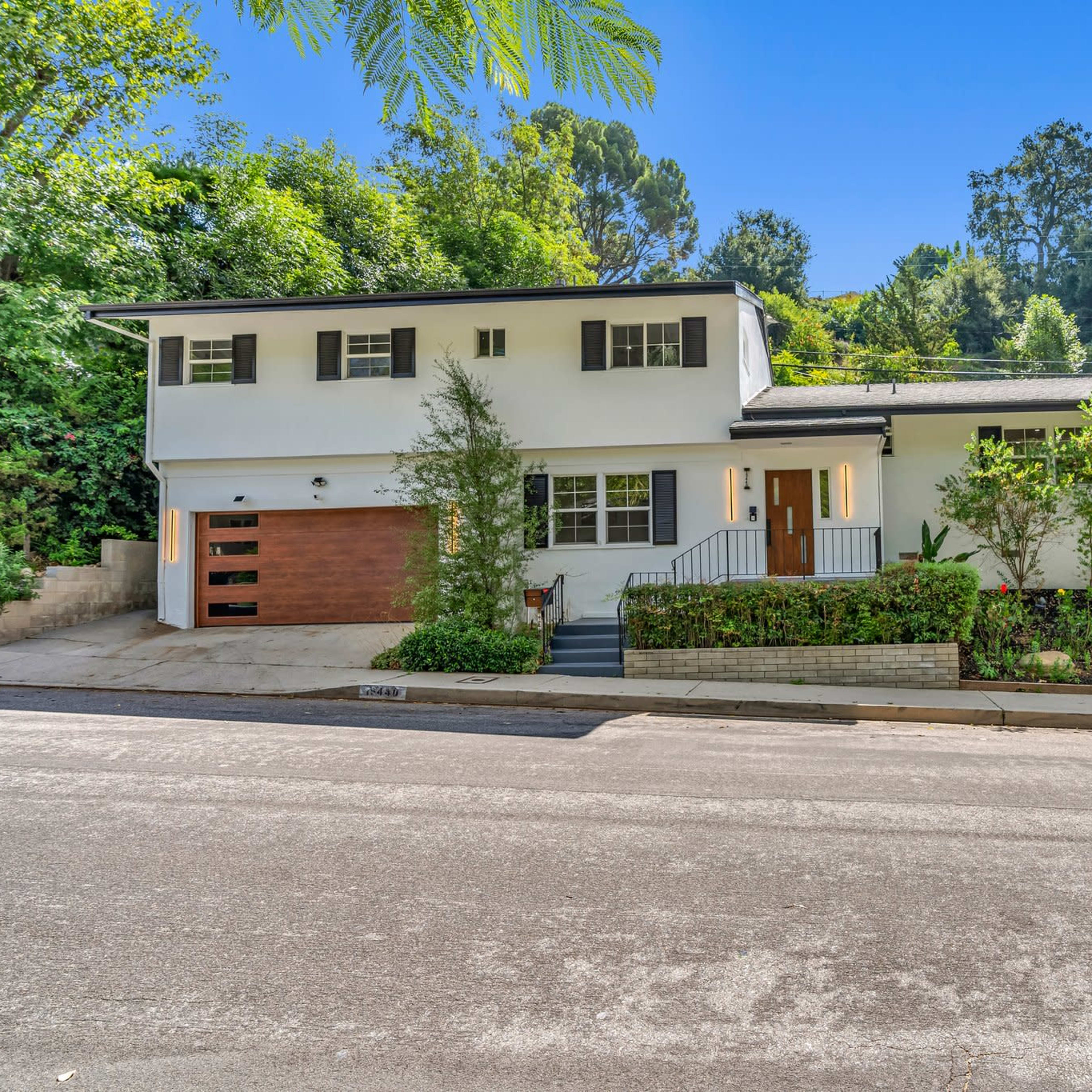 A two-story white house with a brown garage door is surrounded by greenery along a quiet street.