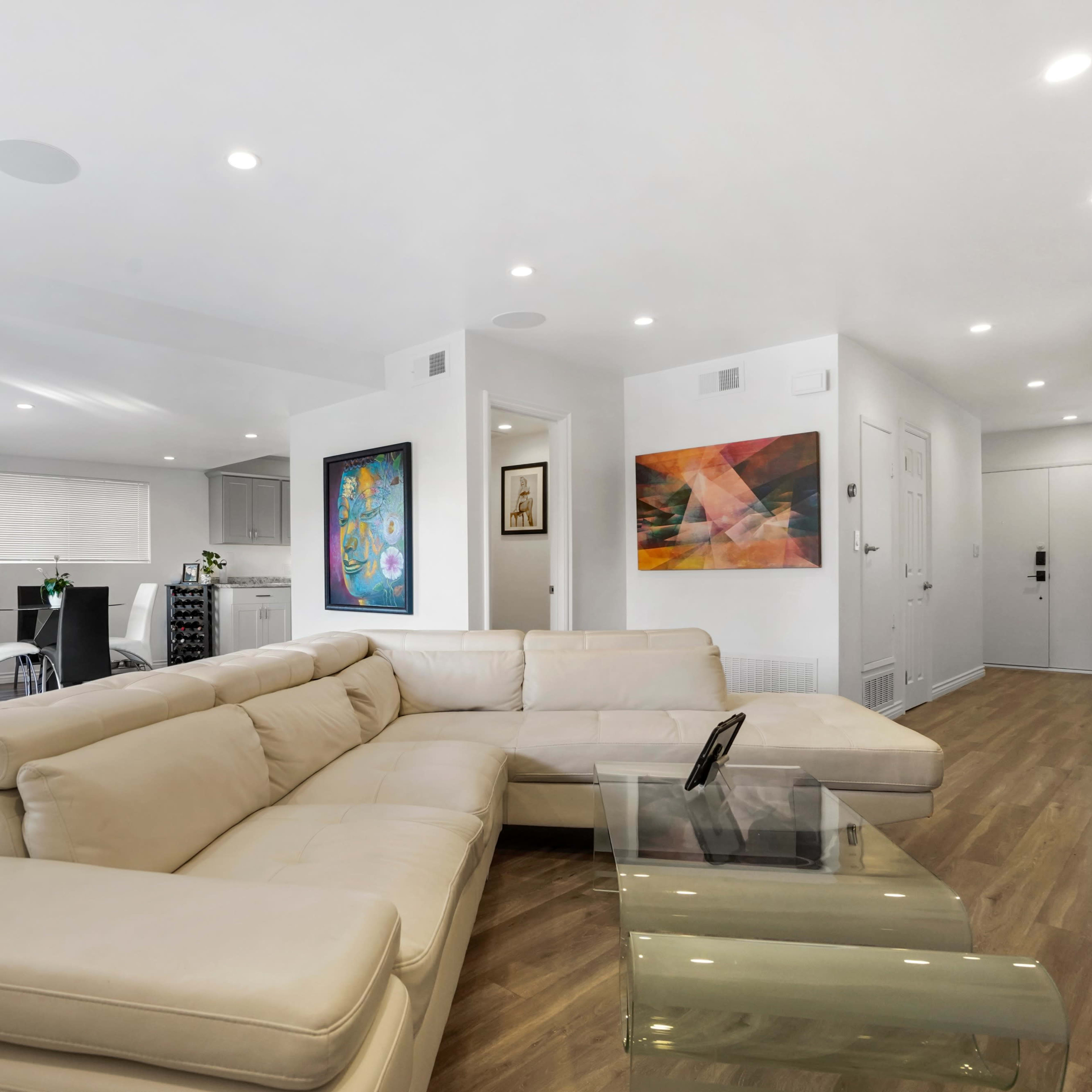 A modern living room with a beige sectional sofa, colorful abstract artwork on the walls, and an open layout leading to a dining area.