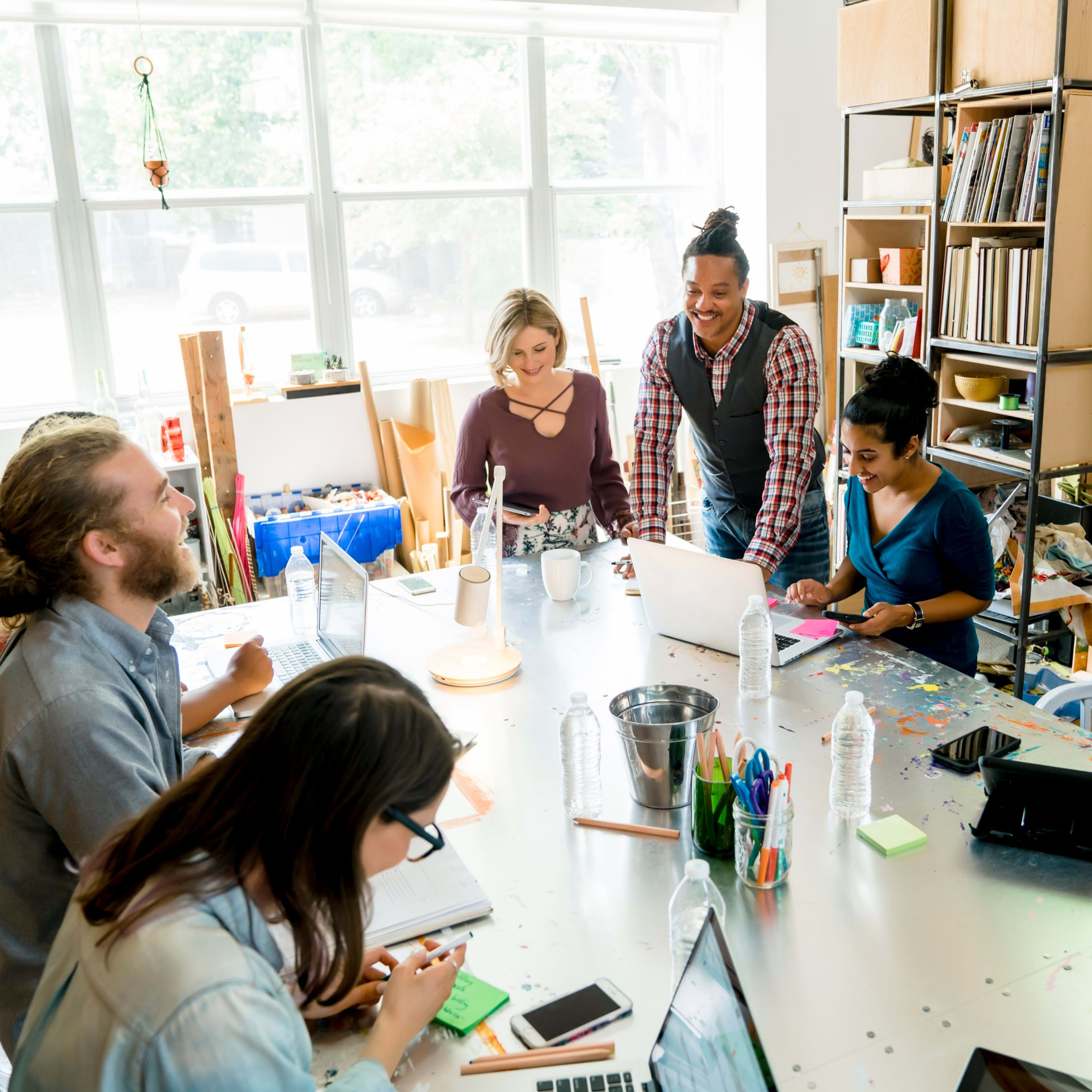 A group of six people collaborates around a large table filled with notebooks and laptops in a bright, well-organized studio.