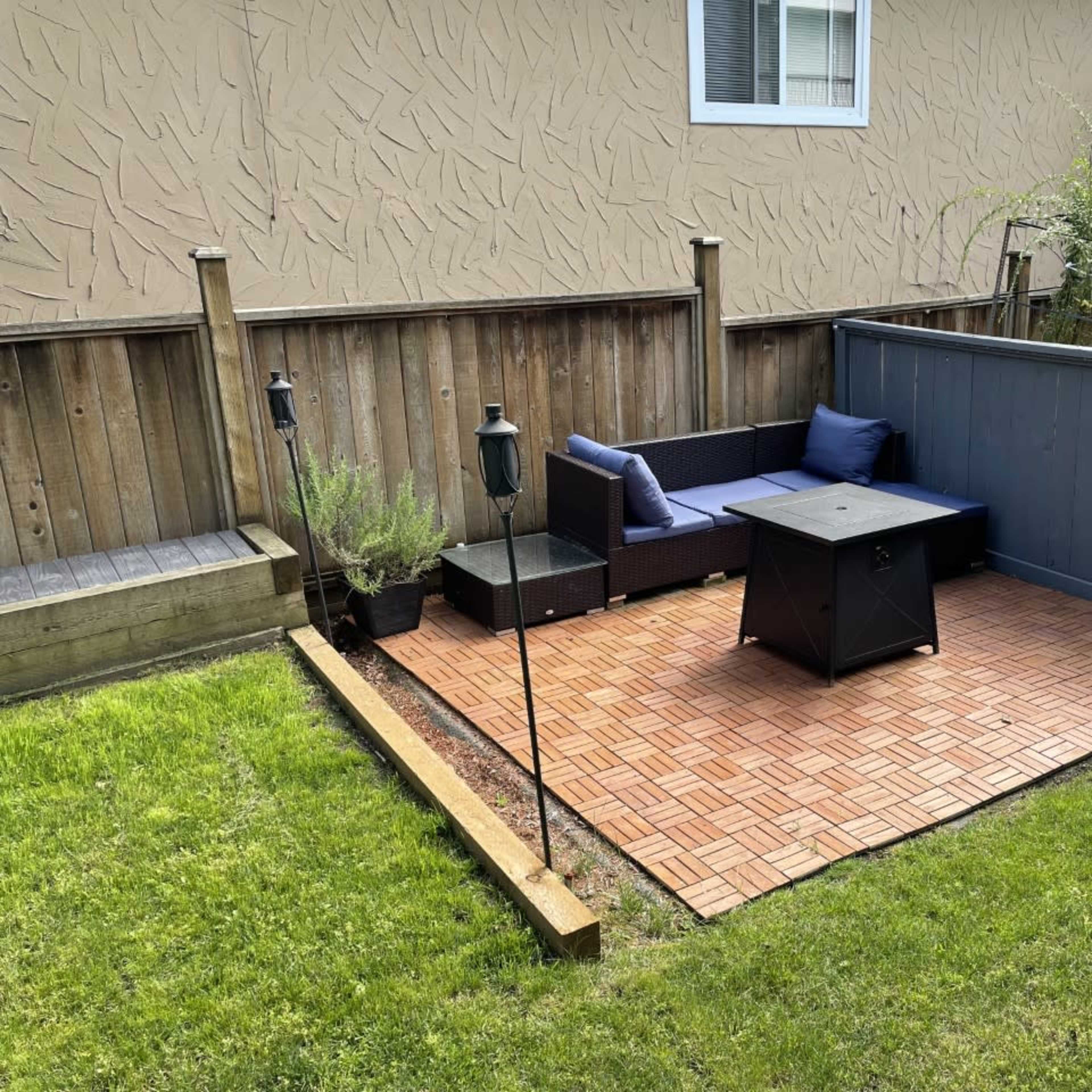 A small outdoor seating area with a black table and a sectional couch, surrounded by grass and a wooden fence.