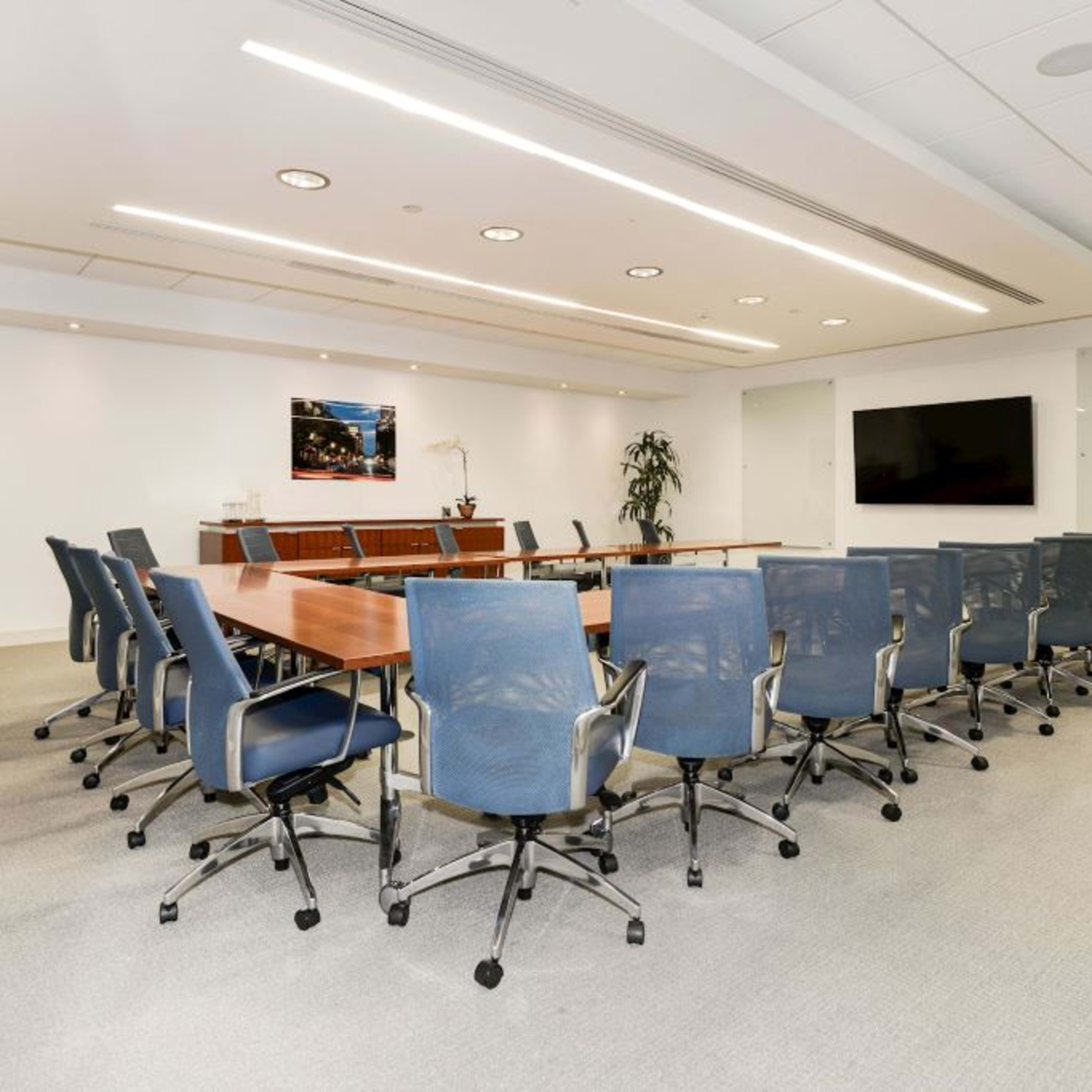 A modern conference room features a long wooden table surrounded by ergonomic chairs, with a large screen and potted plants in the background.