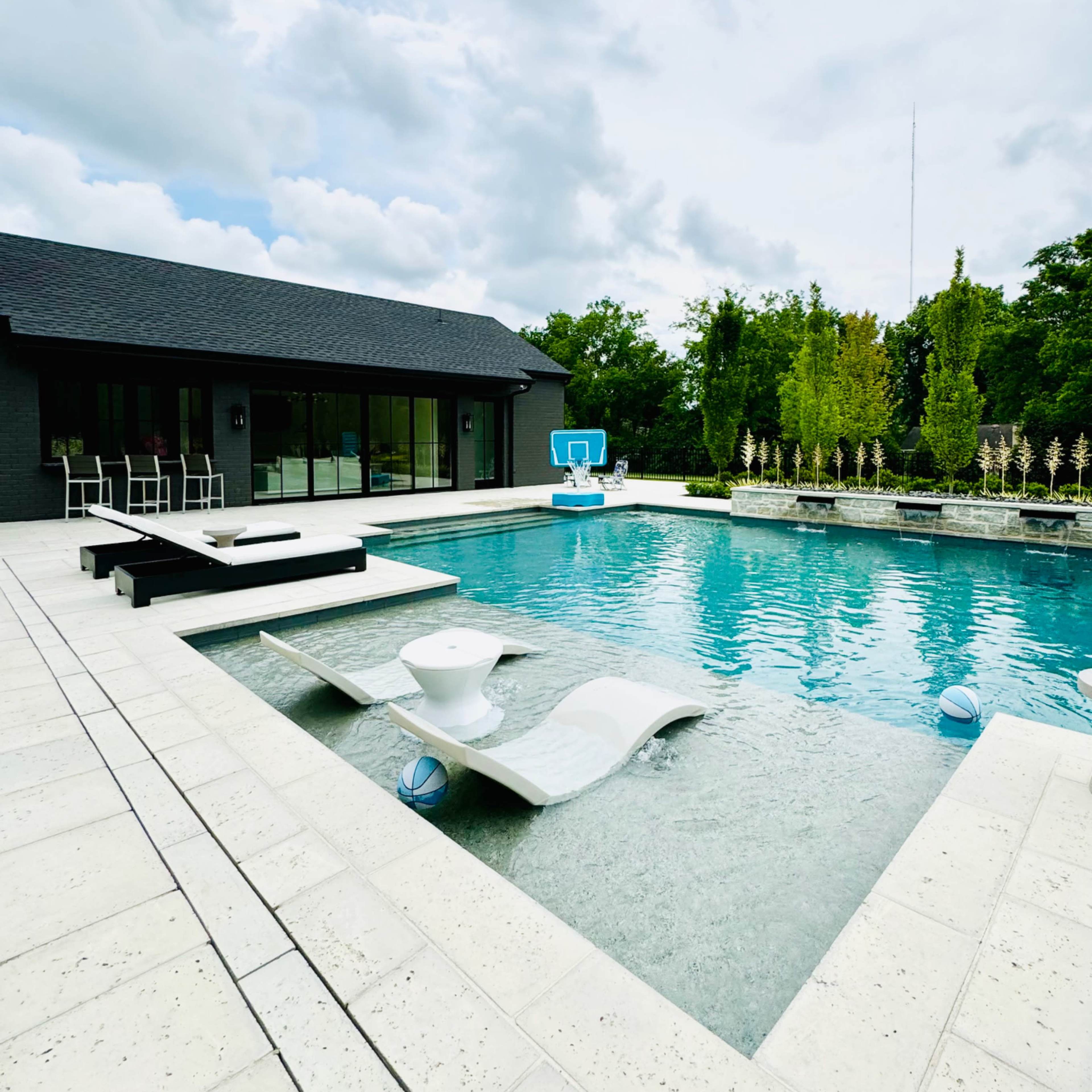A modern swimming pool surrounded by a patio and a contemporary house in a landscaped outdoor area under a cloudy sky.