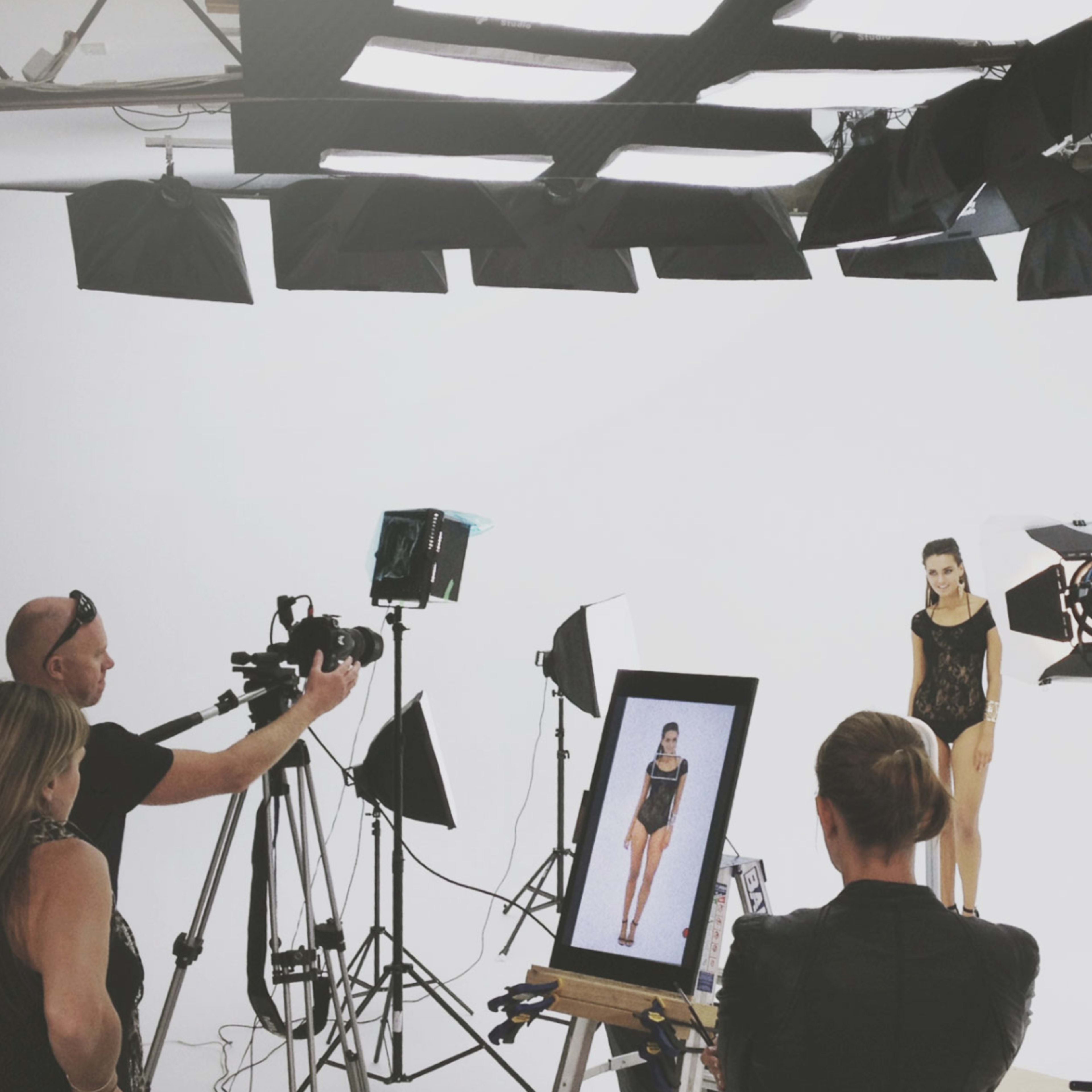 A modeling photoshoot is taking place in a studio with multiple cameras and lighting setups focusing on a model in front of a white background.