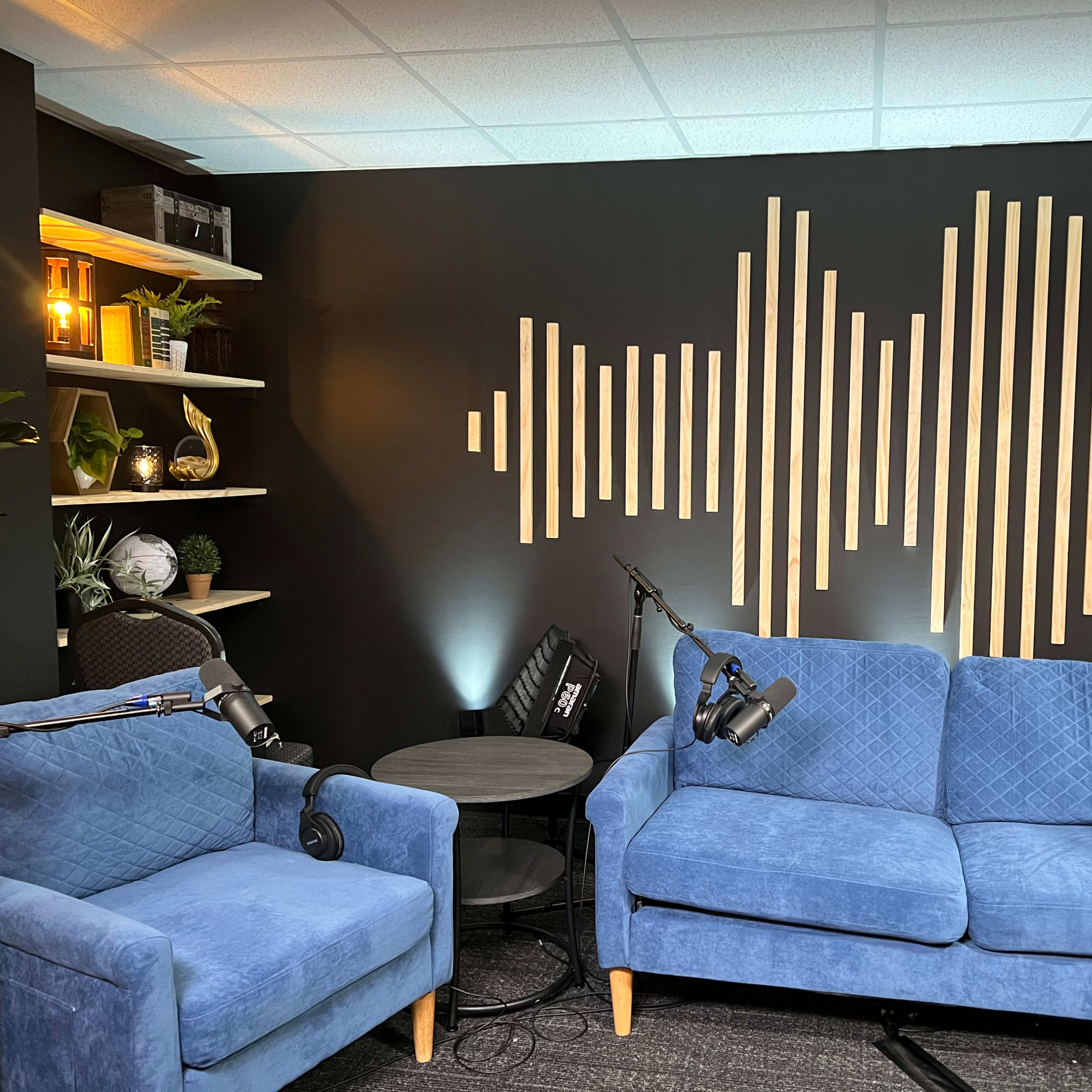 A recording studio is set up with two blue couches, a small black table, and microphones positioned on stands, against a wall featuring a wooden soundwave design.