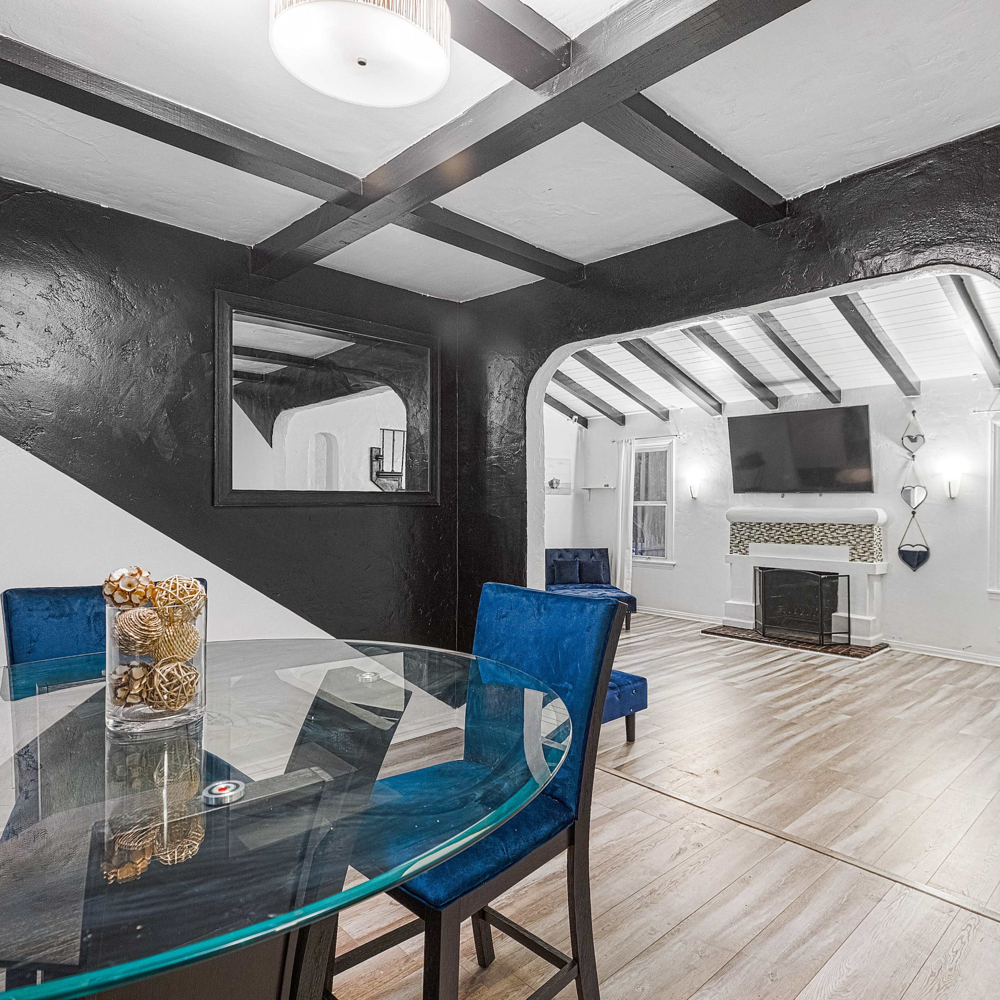 A dining area with a glass table and blue chairs leads into a living room featuring a fireplace and a television on the wall.