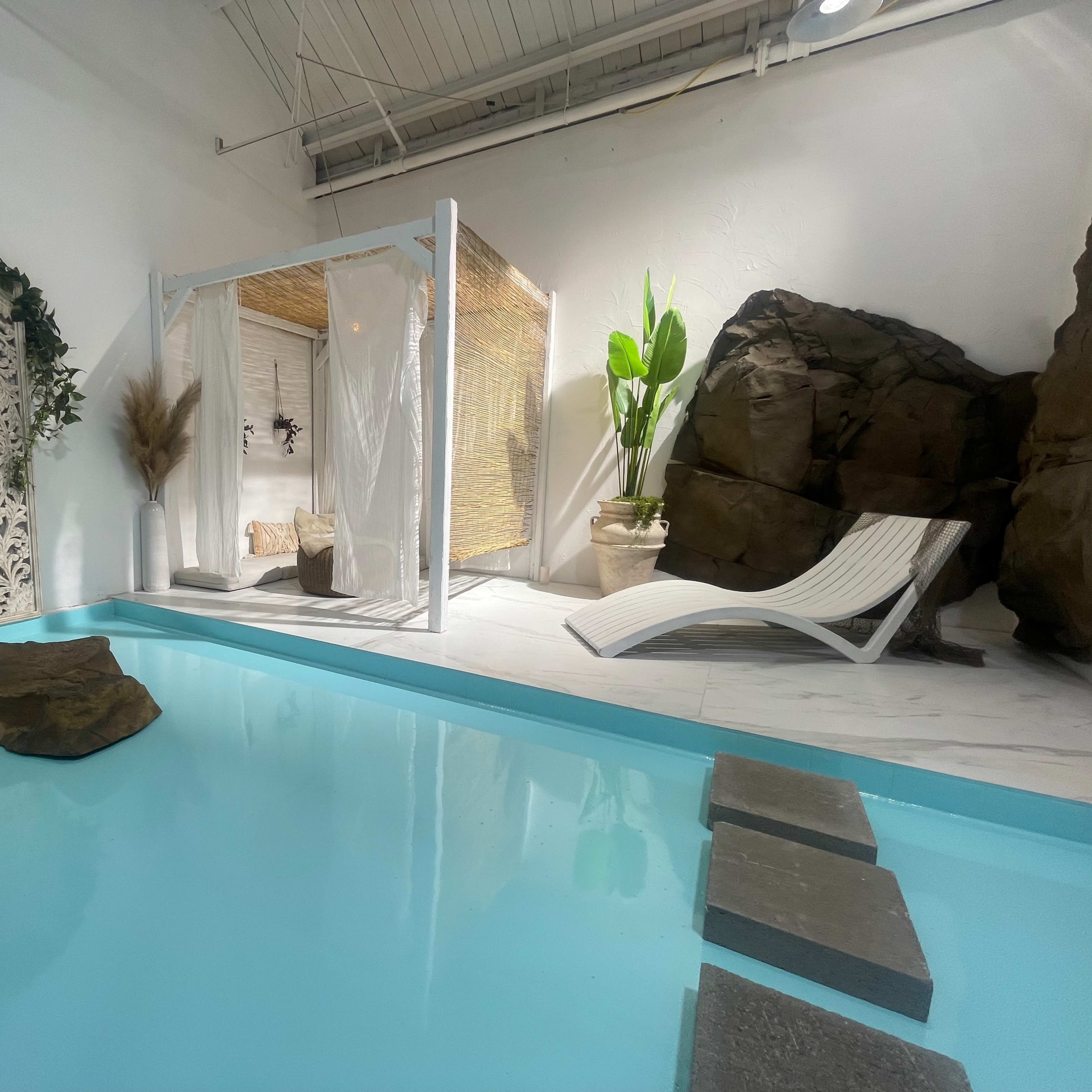 A modern indoor space features a shallow turquoise pool surrounded by decorative rocks, a sun lounger, and a screened area with plants and artwork.