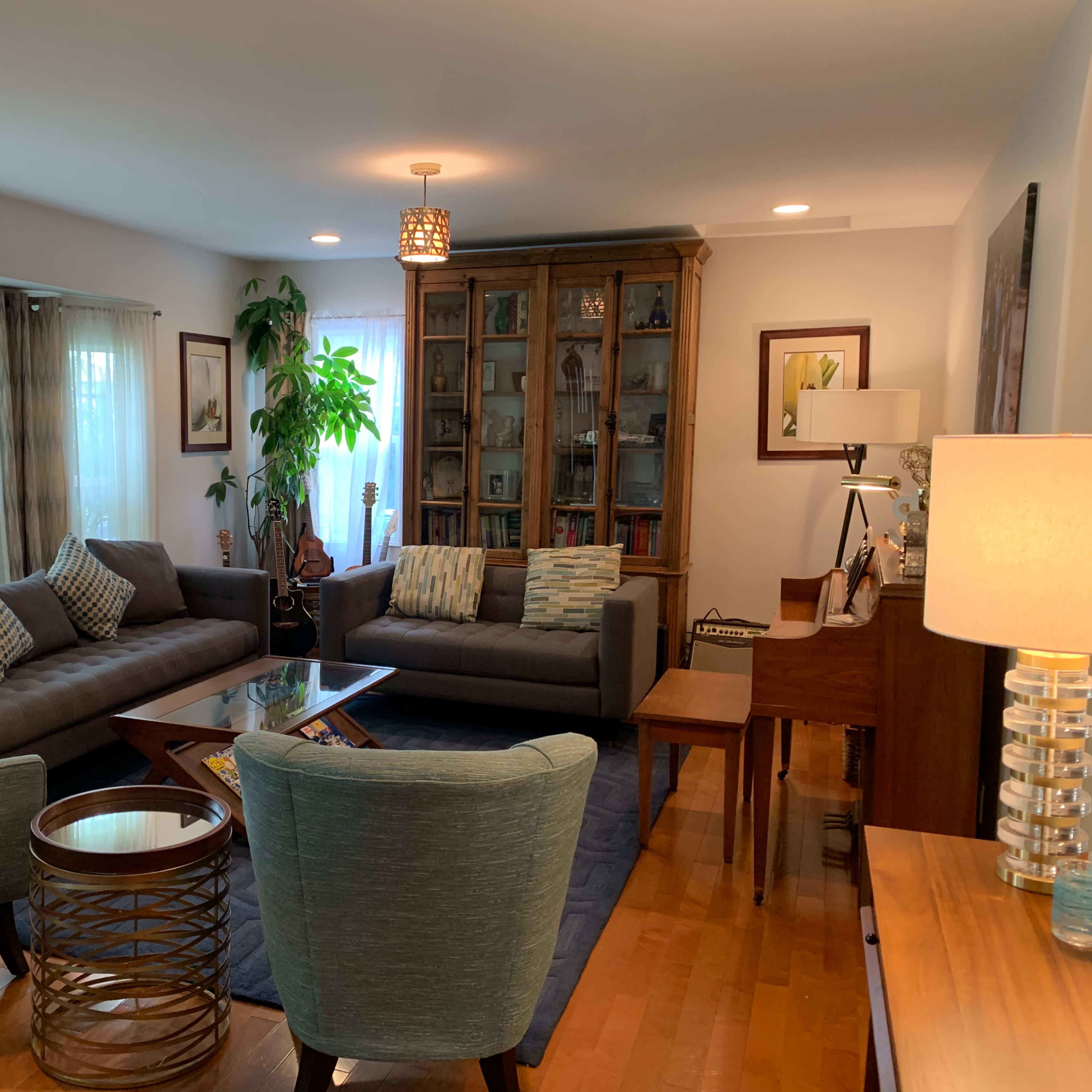 A modern living room features two gray sofas, a glass coffee table, a wooden bookshelf, and a piano, with natural light streaming through the windows.
