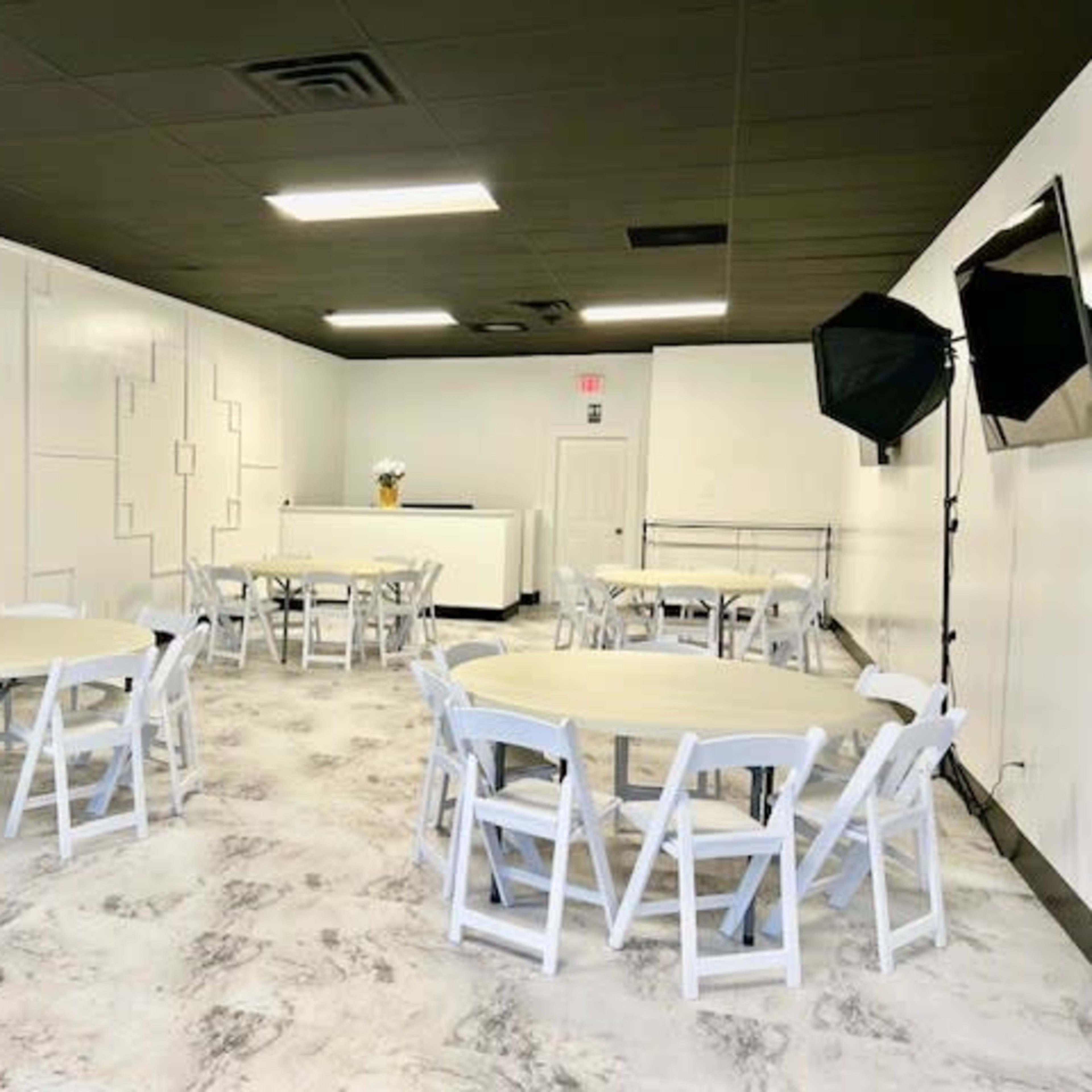 A simple, empty event space with white walls, several round tables with white chairs, and a small stage area at the front.