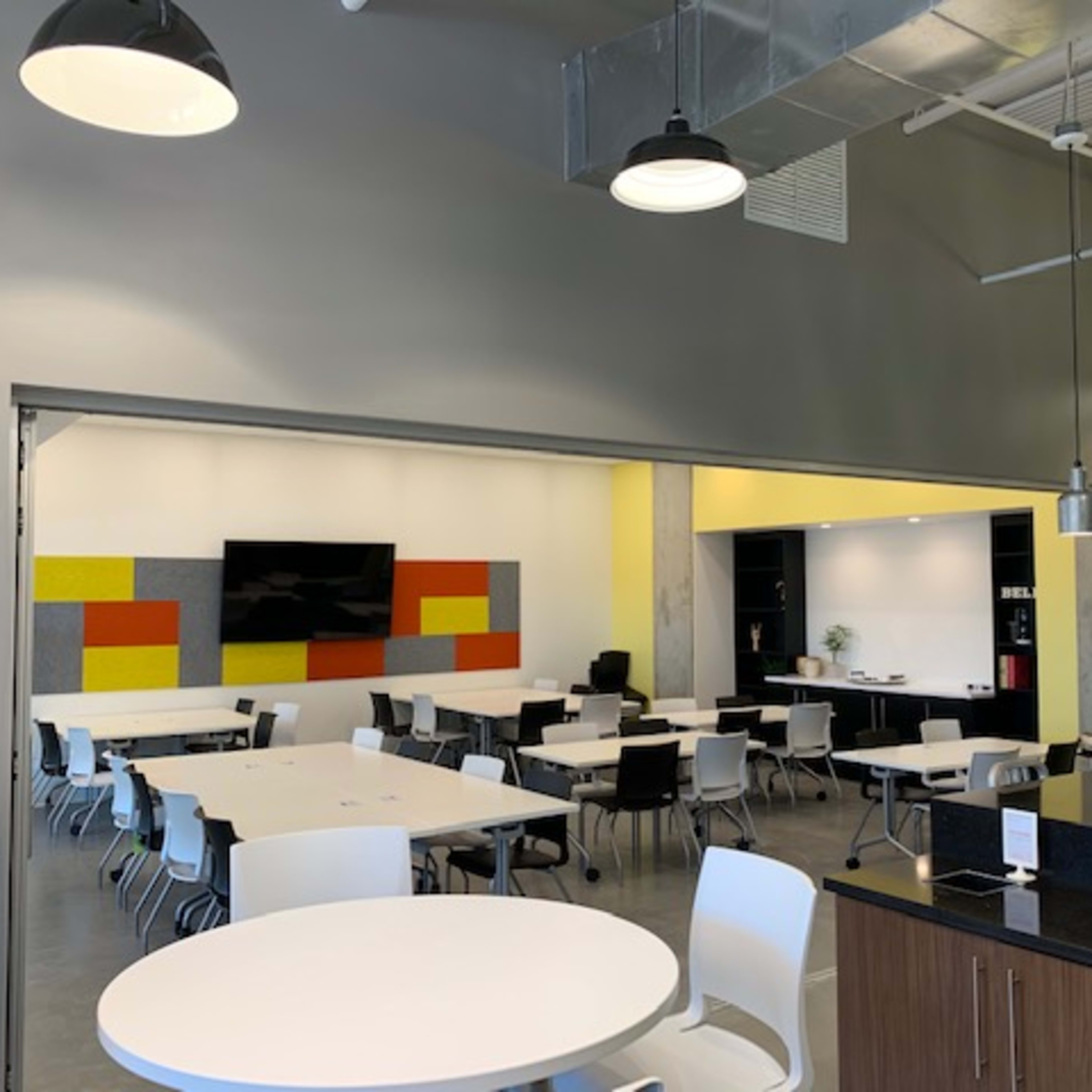 A modern, open-plan dining area with white tables and chairs, a large television screen on the wall, and a neighboring workspace featuring shelves and plants.