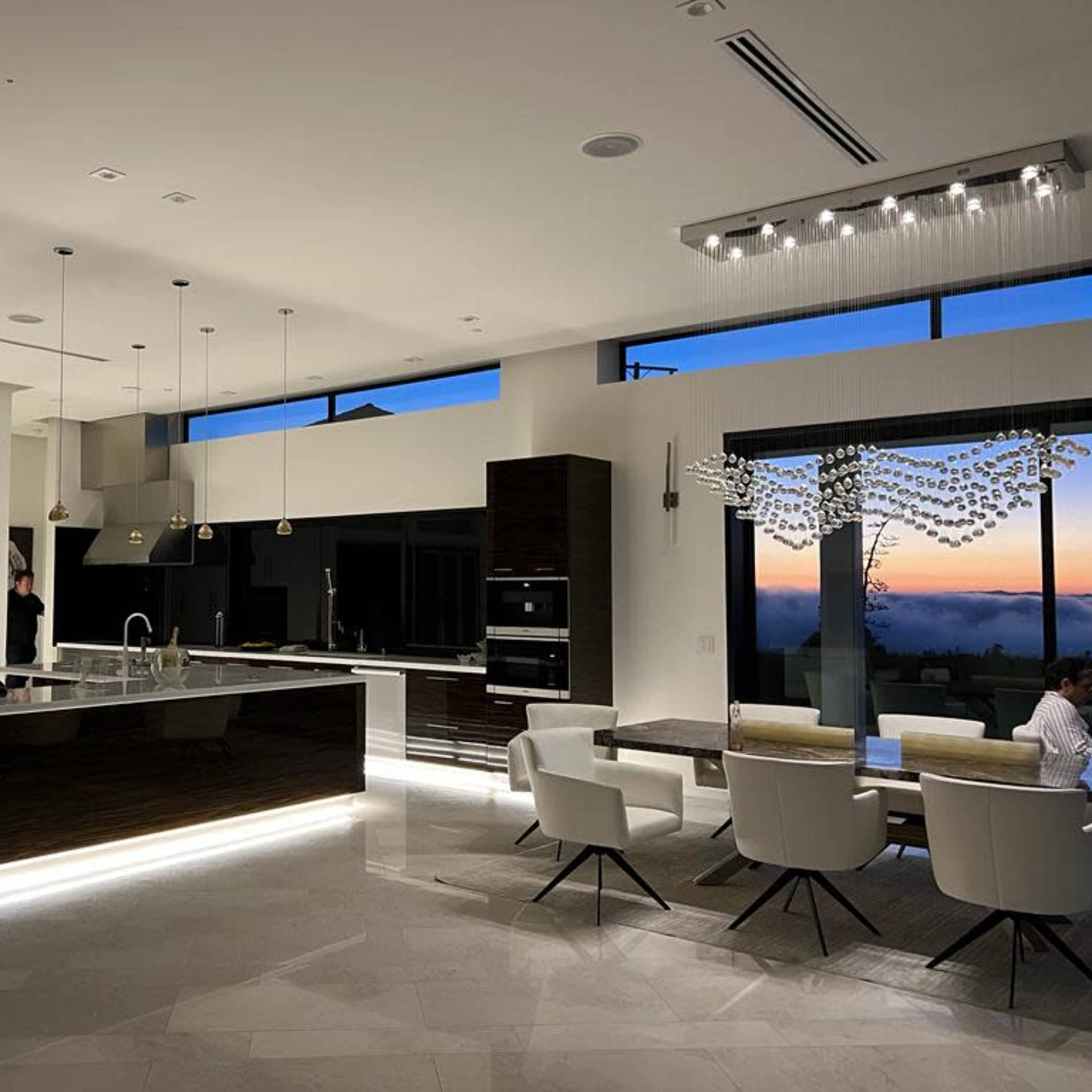 A modern kitchen with a central island, dining area, and large windows displaying a sunset.