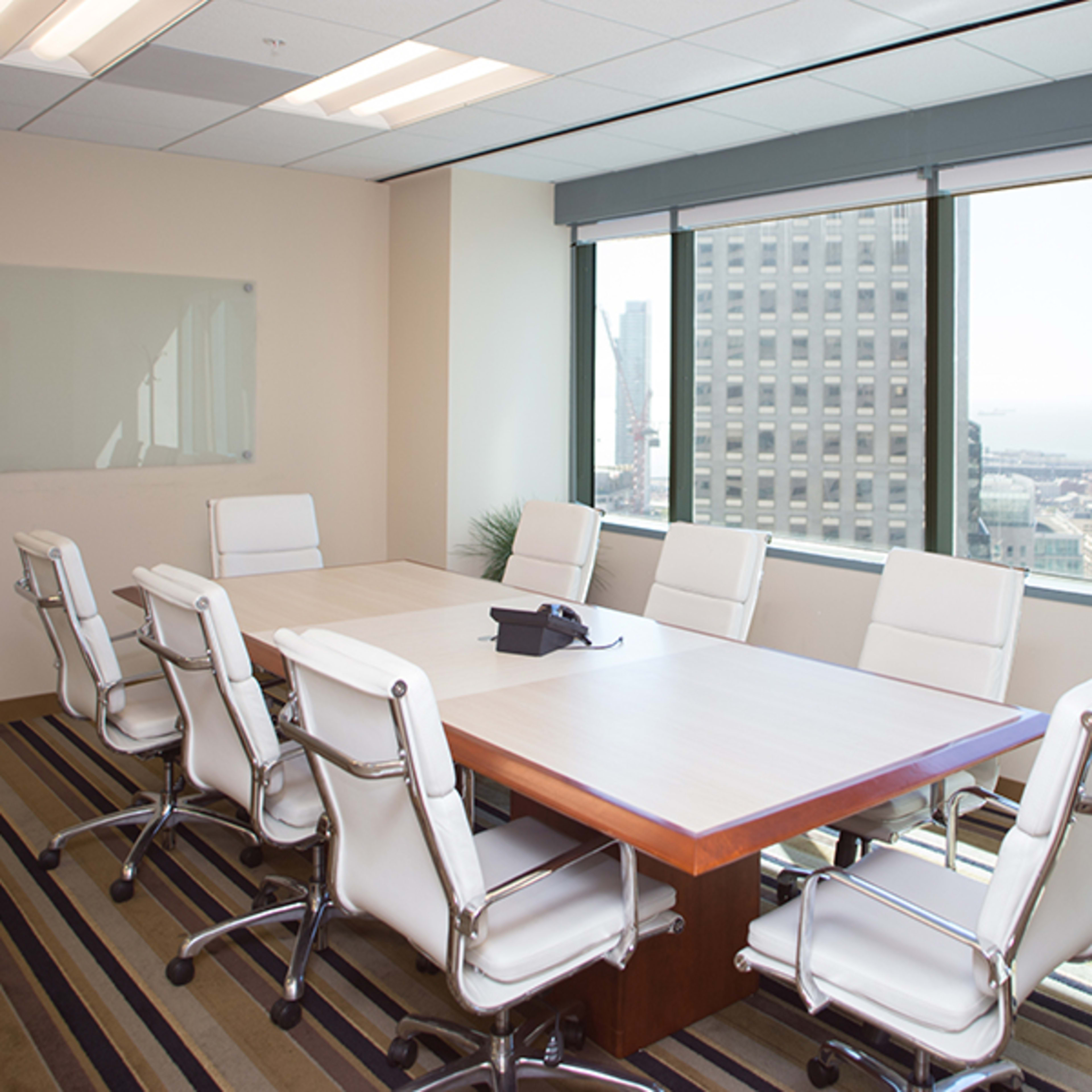 A modern conference room features a large wooden table surrounded by eight white ergonomic chairs and has windows providing a view of the city skyline.