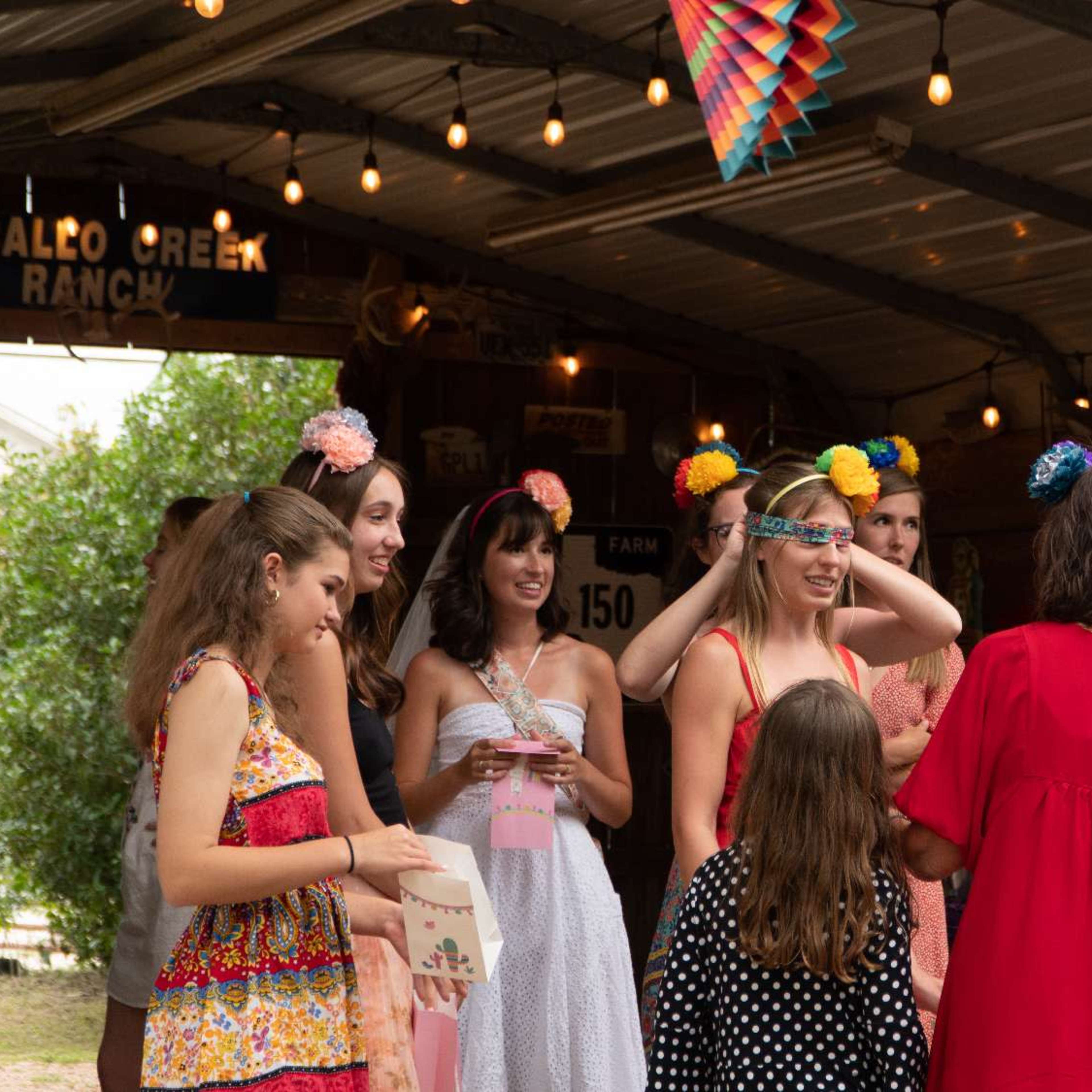 A group of women and girls gather under a decorated shelter at a ranch, with festive decorations and string lights overhead.