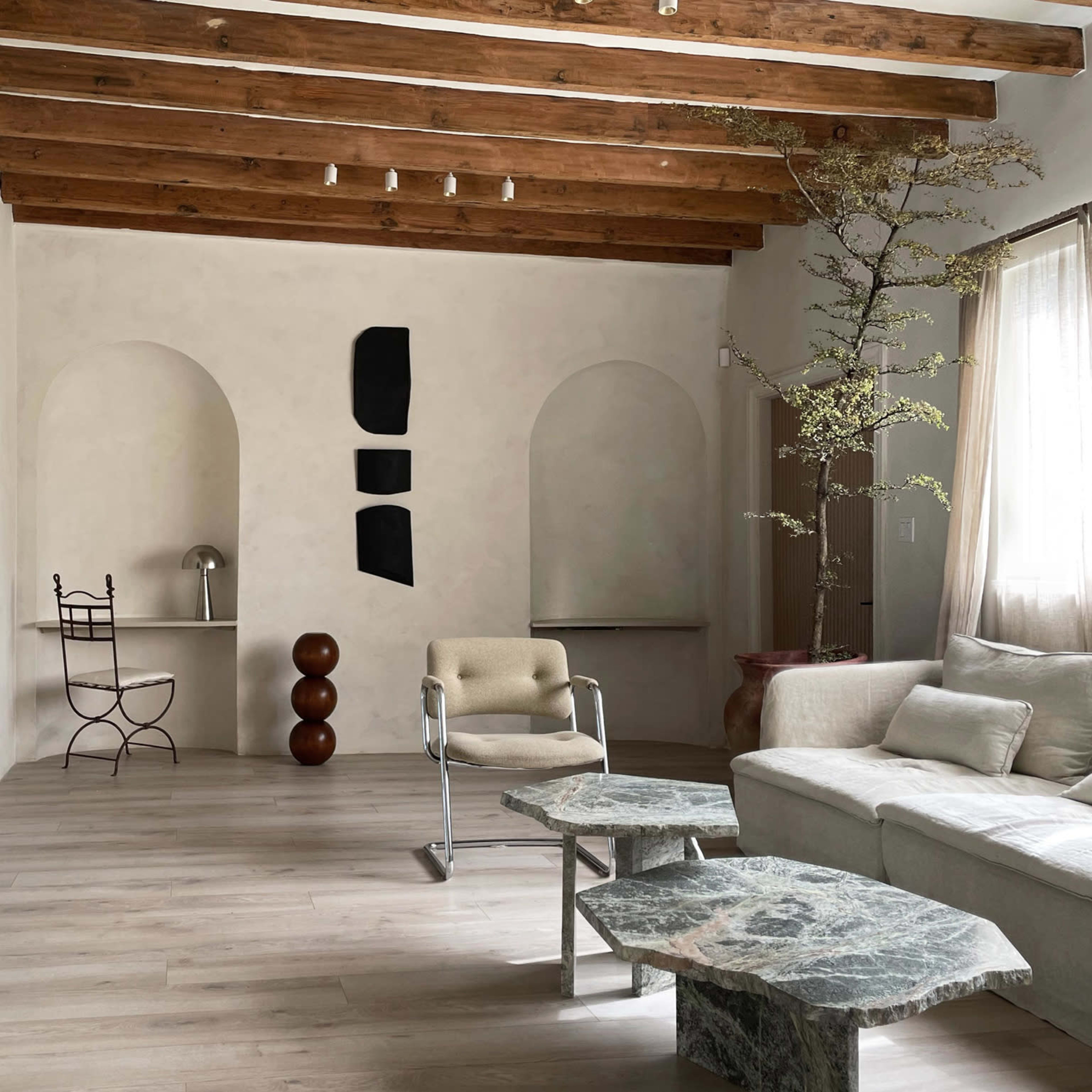 The room features a minimalist design with wooden beams, a beige sofa, a round stone table, and a vertical arrangement of artworks on the wall.