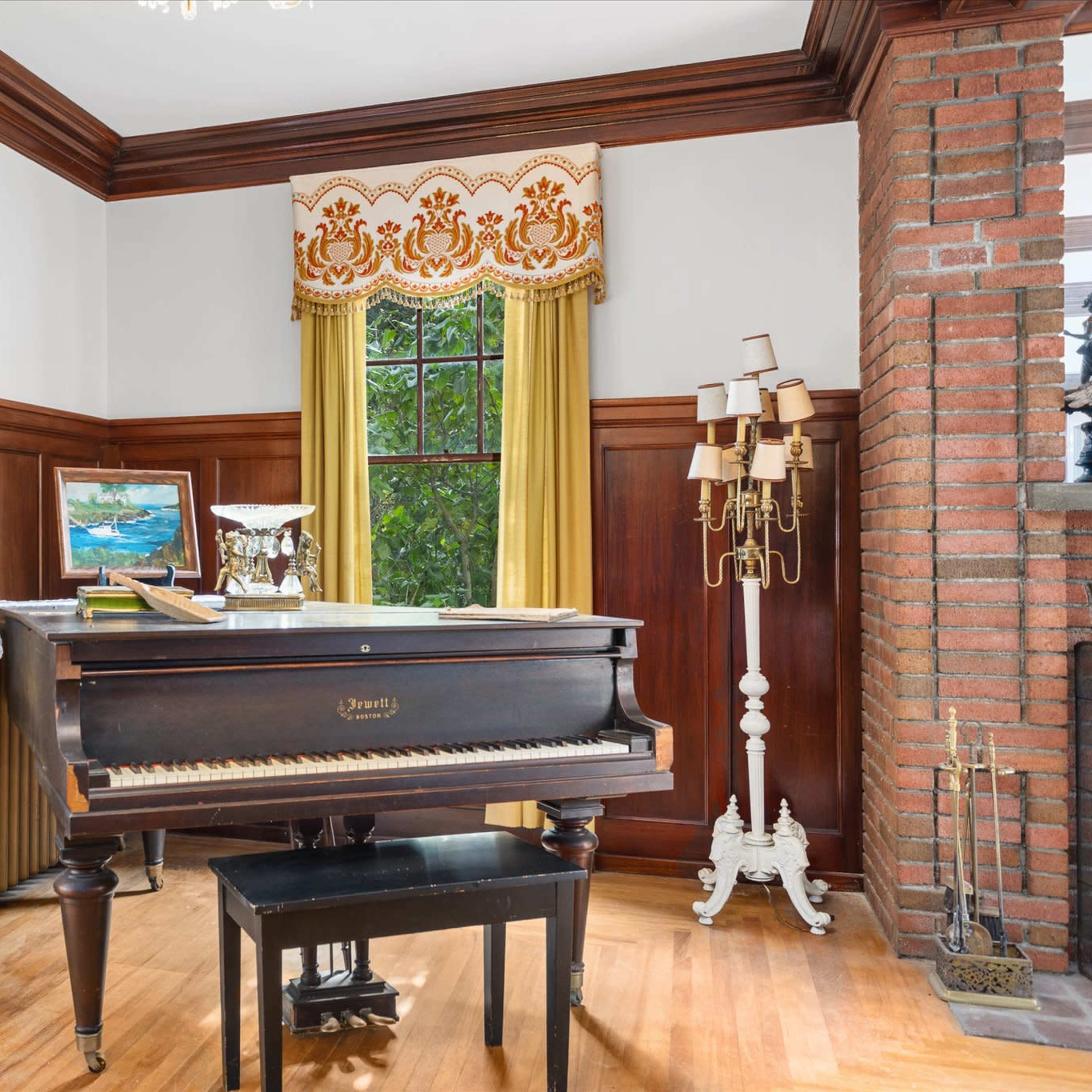 A vintage piano positioned in a room with wooden paneling, a window draped with yellow curtains, and various decorative elements.
