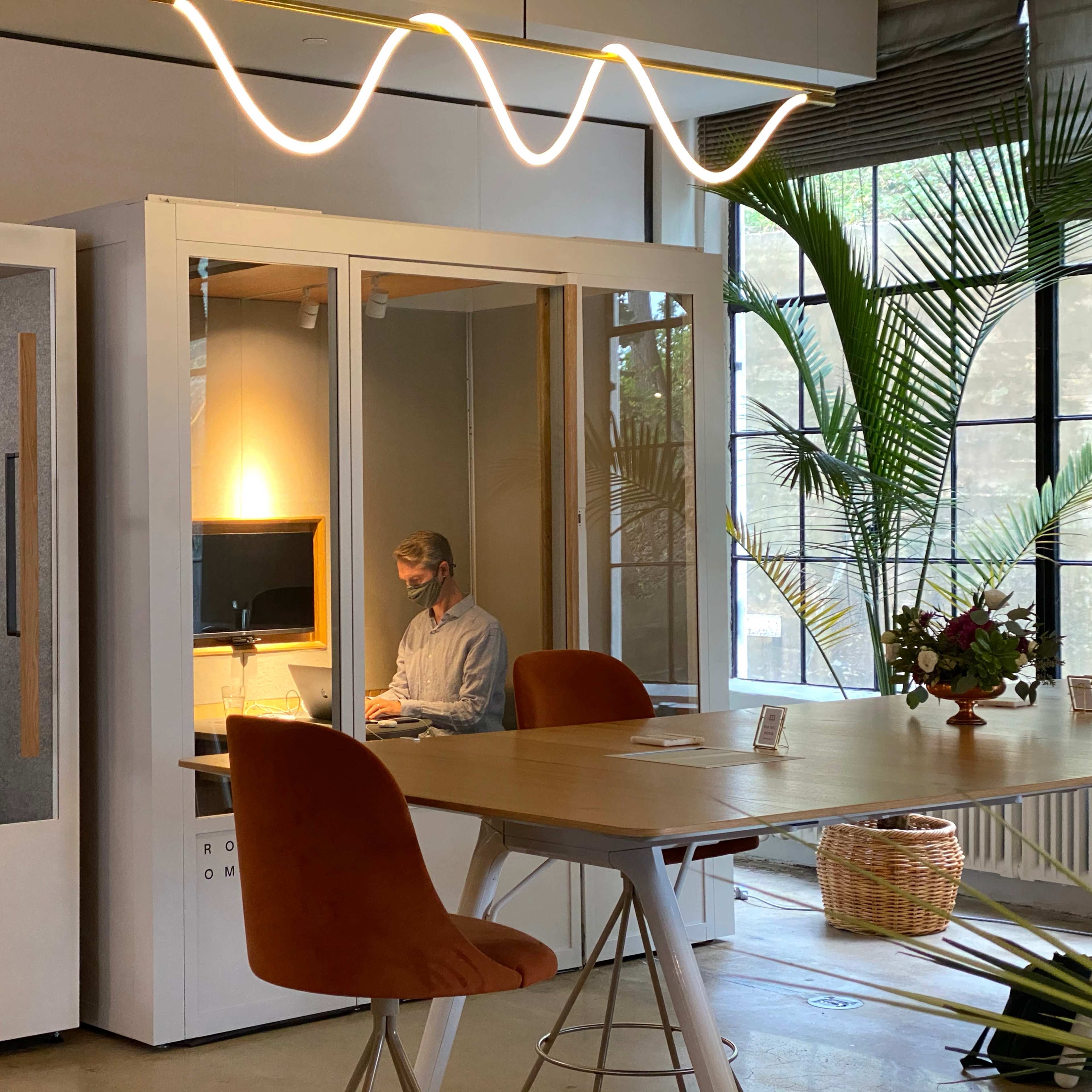 A man works at a desk inside a glass-enclosed workspace while another person uses a nearby booth in a modern office setting.