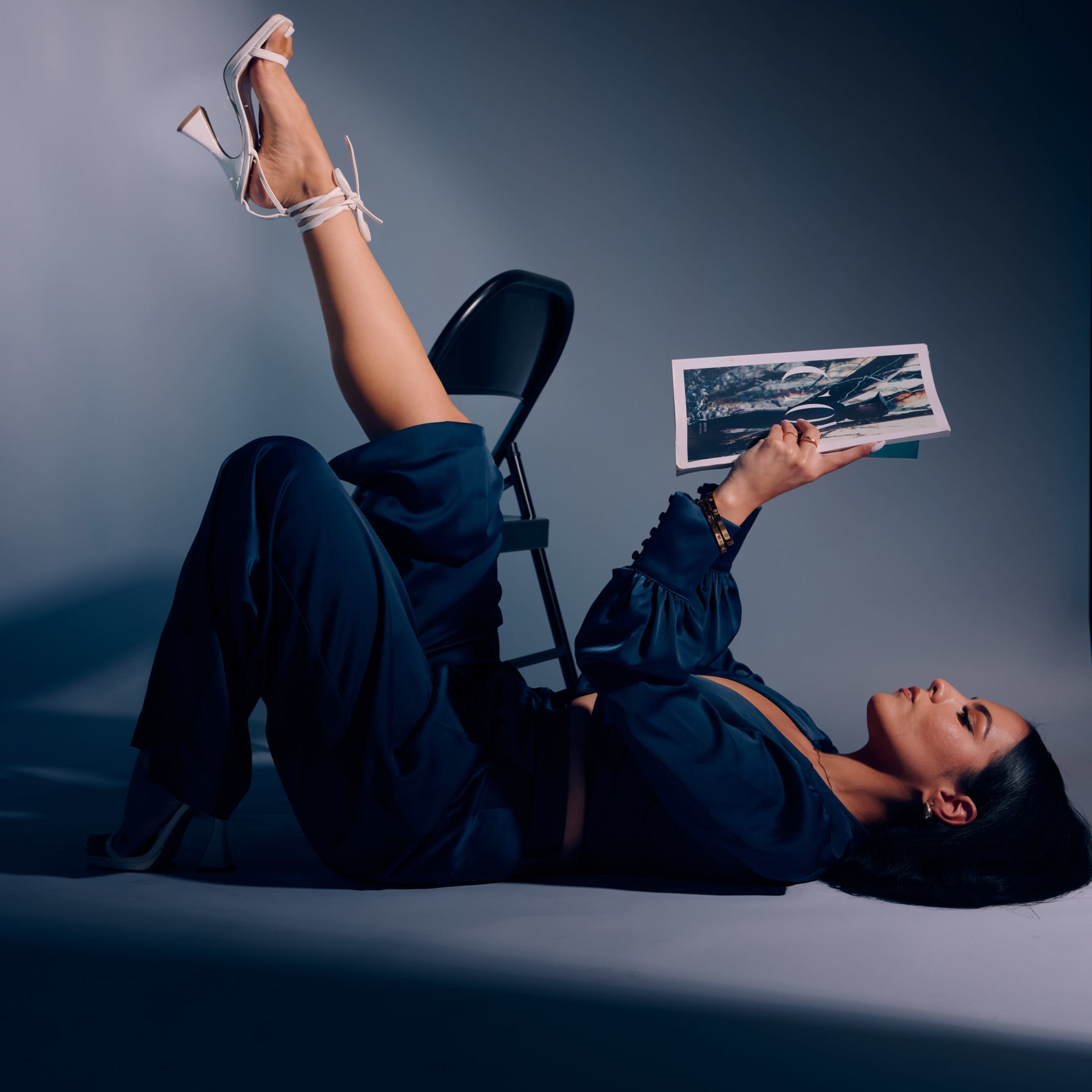 A person lies on the floor with a raised leg, holding a black-and-white photograph, while resting against a chair in a dimly lit studio.