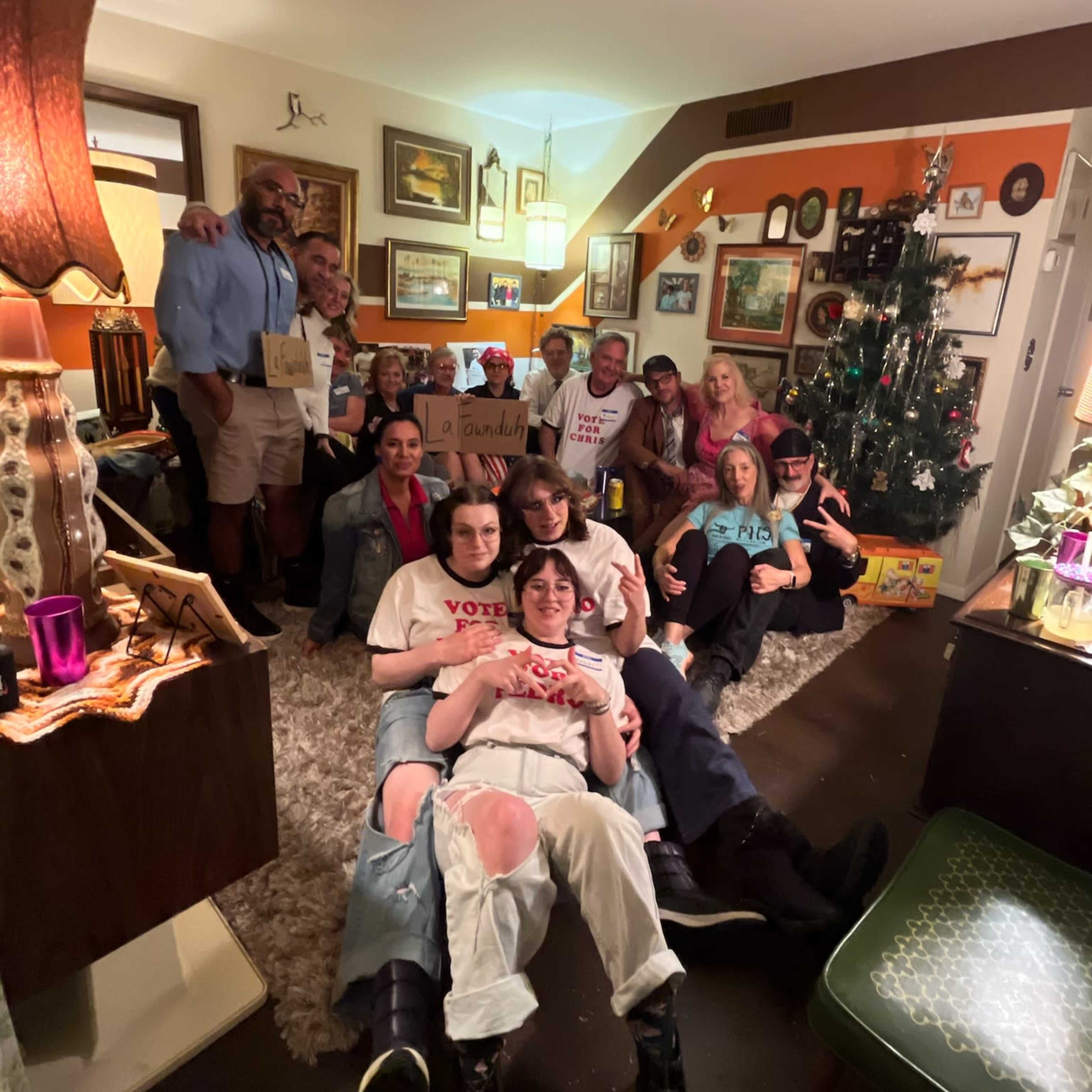 A diverse group of people is gathered in a cozy living room, posing for a photo with festive decorations and a Christmas tree in the background.
