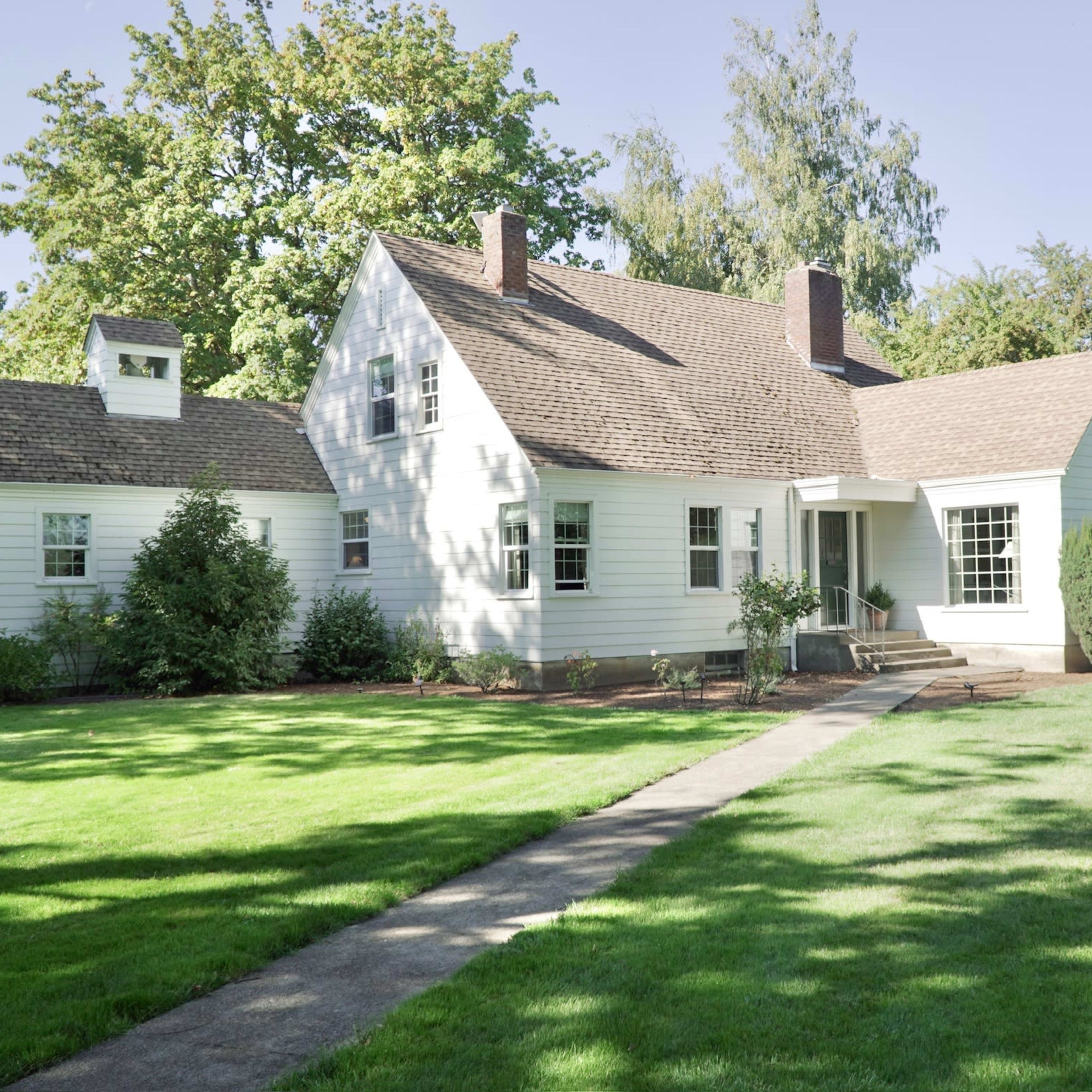 A white, two-story house with a sloped roof, surrounded by green grass and trees, with a concrete pathway leading to the front entrance.