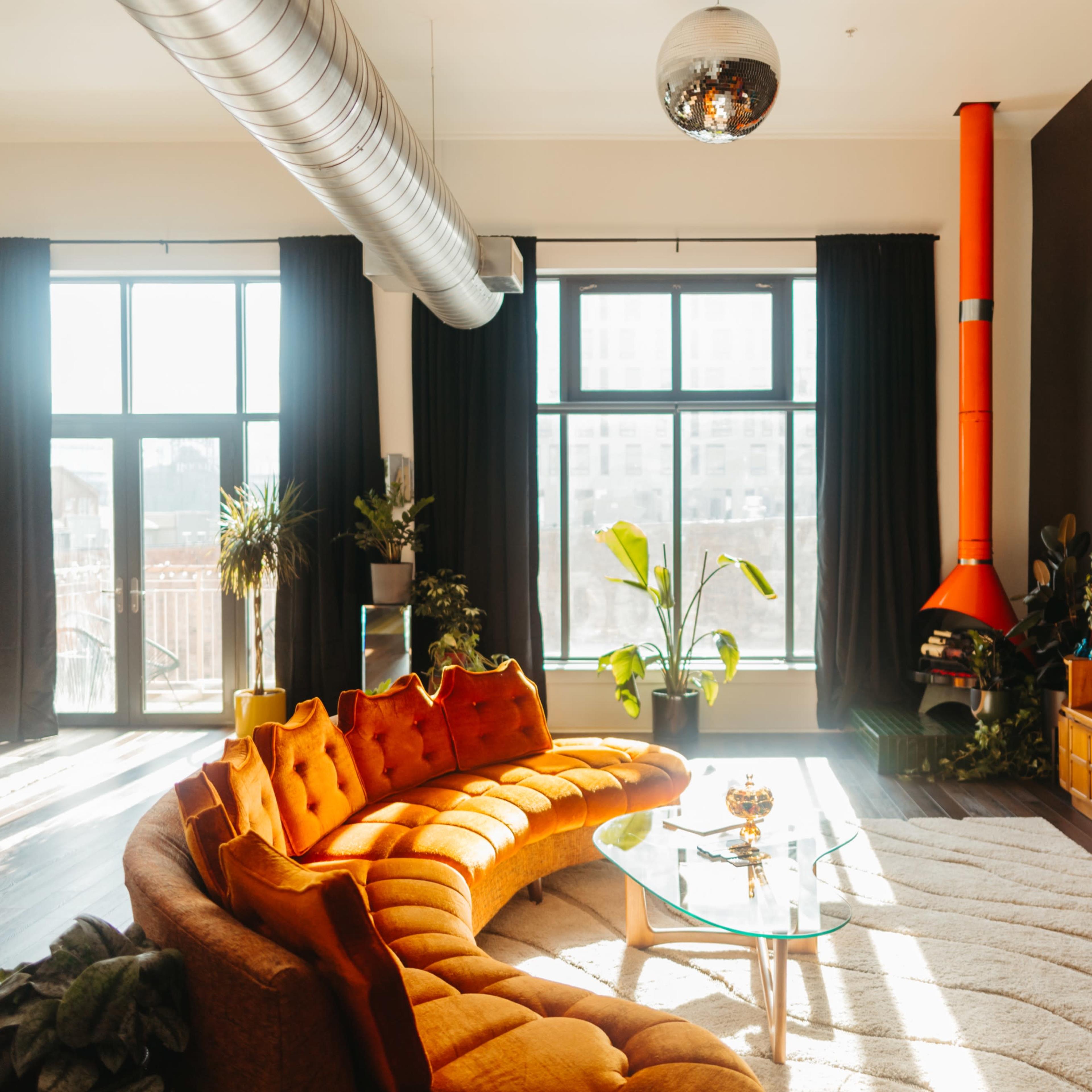 A bright, modern living room features a curved orange sofa, a glass coffee table, large windows with sheer curtains, and a colorful fireplace.
