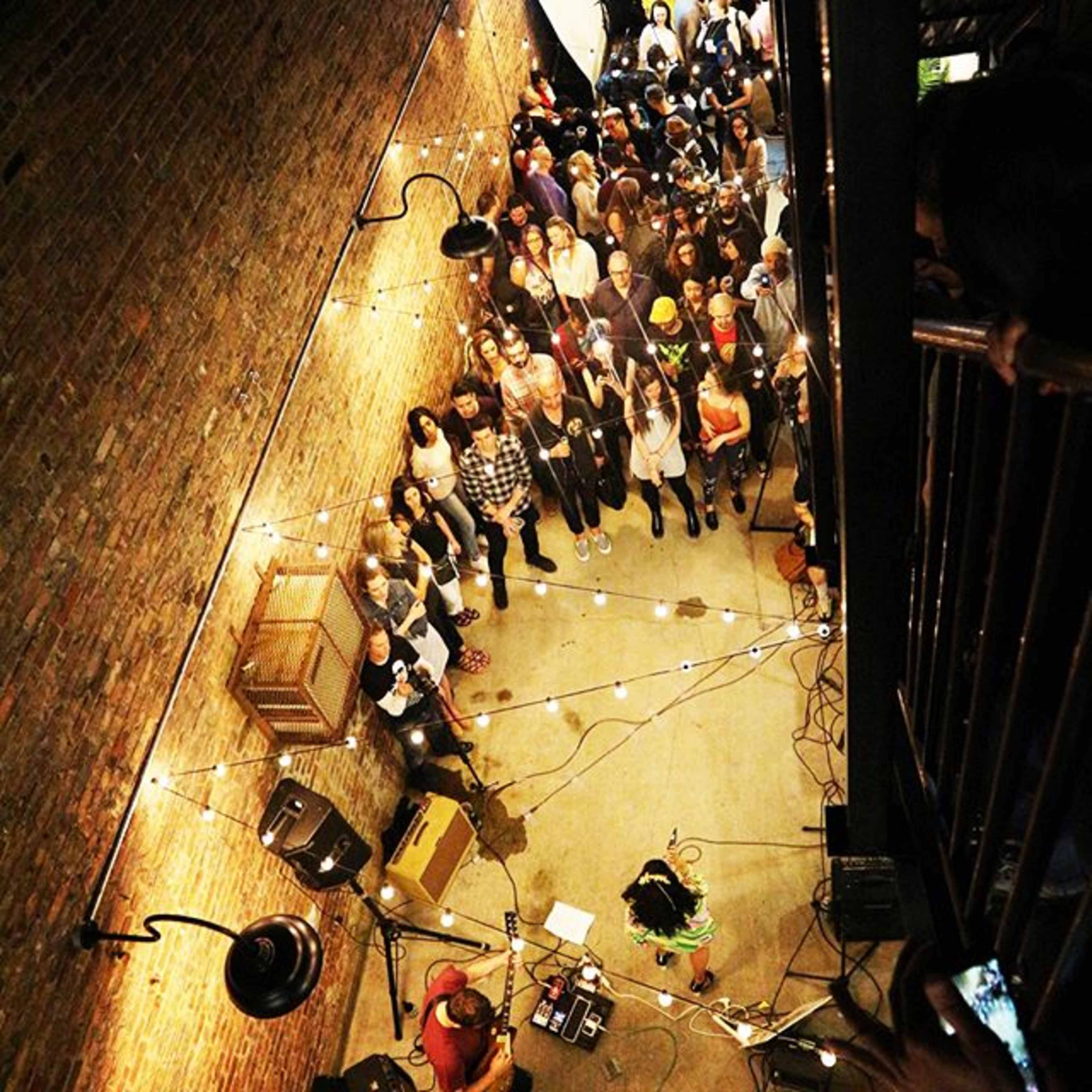 A crowd gathers in a dimly lit venue with exposed brick walls, watching a performer on stage surrounded by string lights.