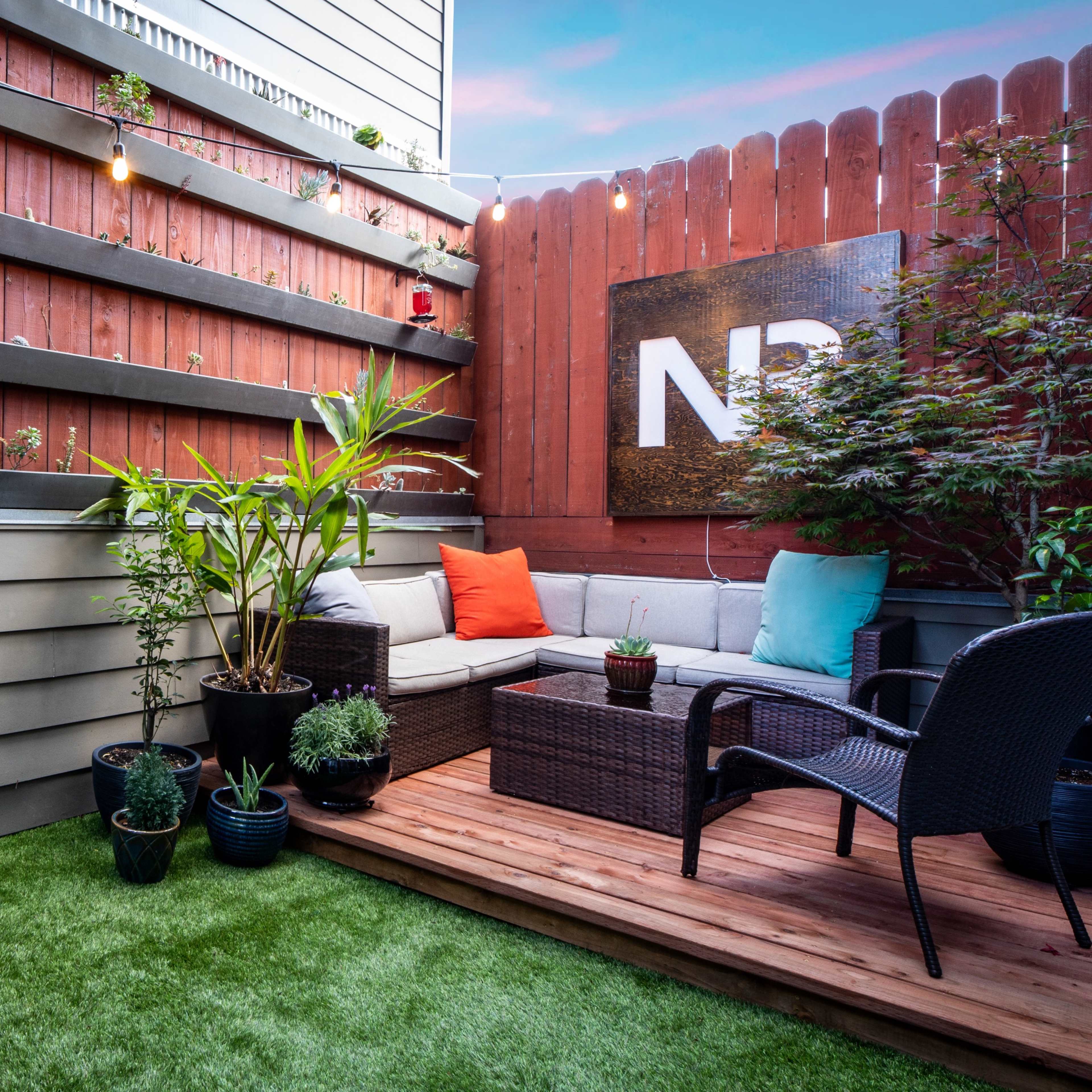 A cozy outdoor patio with a fire pit, seating area, and various potted plants against a wood-paneled wall.