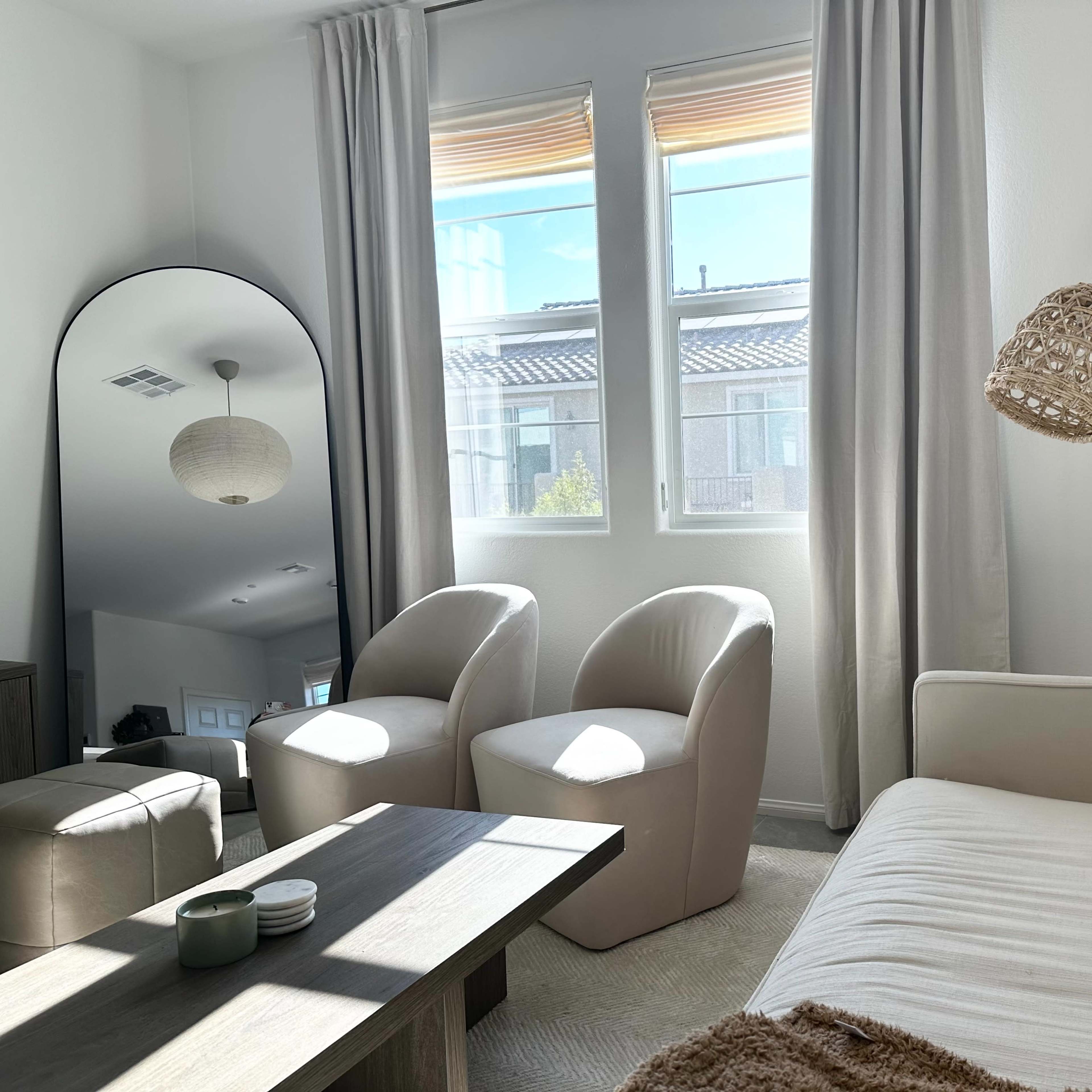 A modern living room with a white sofa, two sculptural chairs, a wooden coffee table, and large windows allowing natural light to illuminate the space.