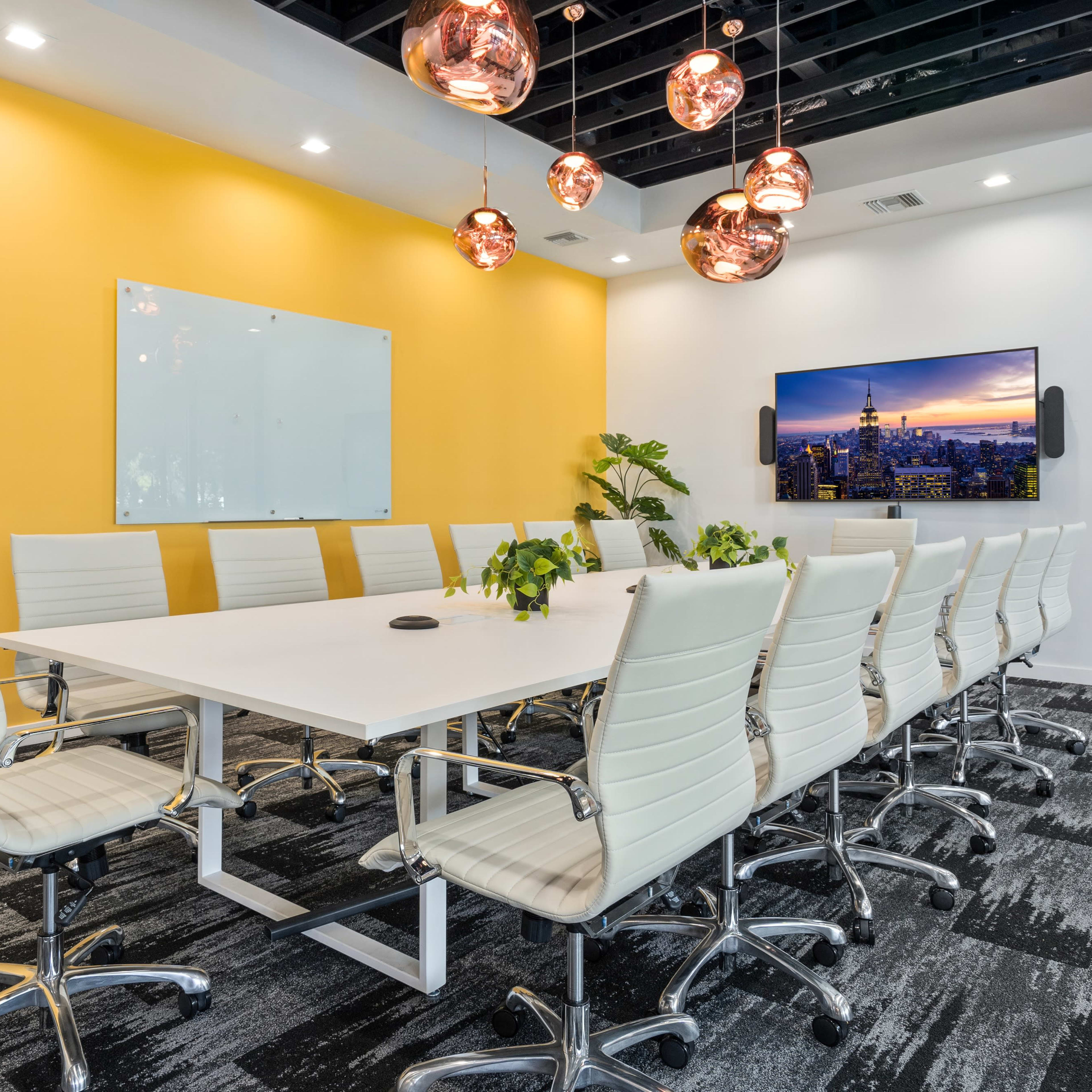 A bright conference room features a large white table surrounded by twelve white chairs, with modern pendant lights hanging above and a wall-mounted screen displaying a city skyline.