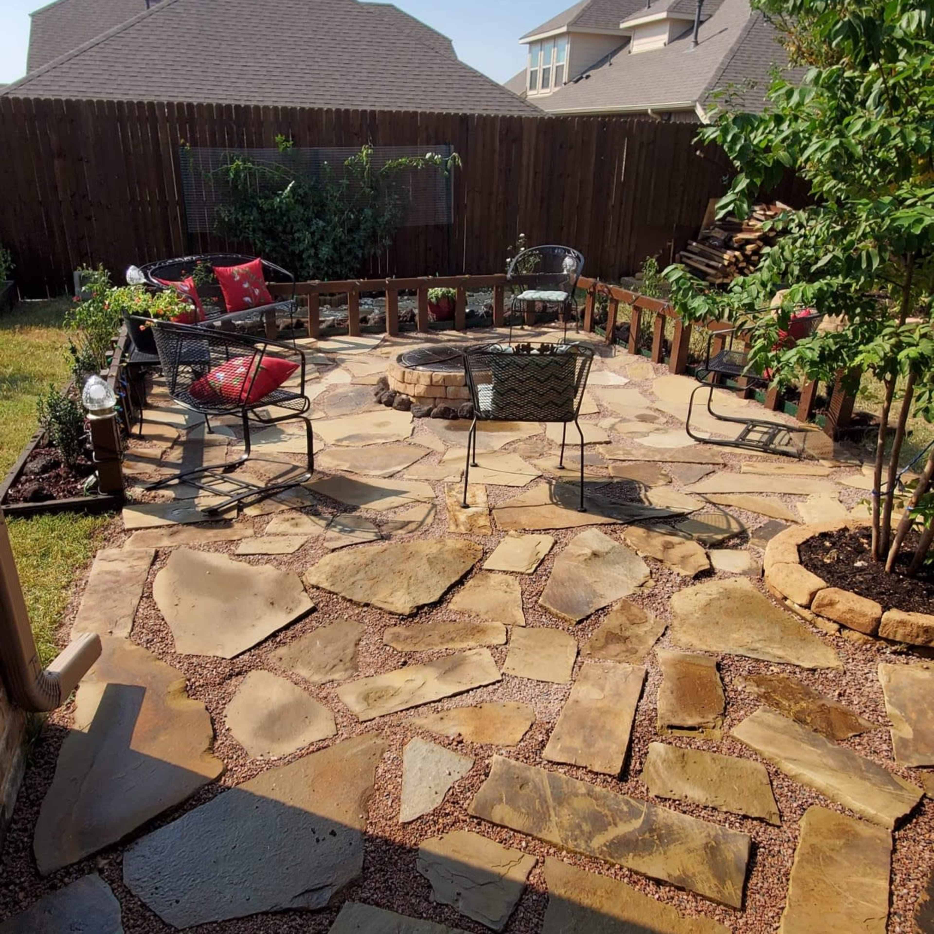 A backyard patio made of large stone slabs surrounded by plants and a fenced area, with several chairs and a table arranged for seating.