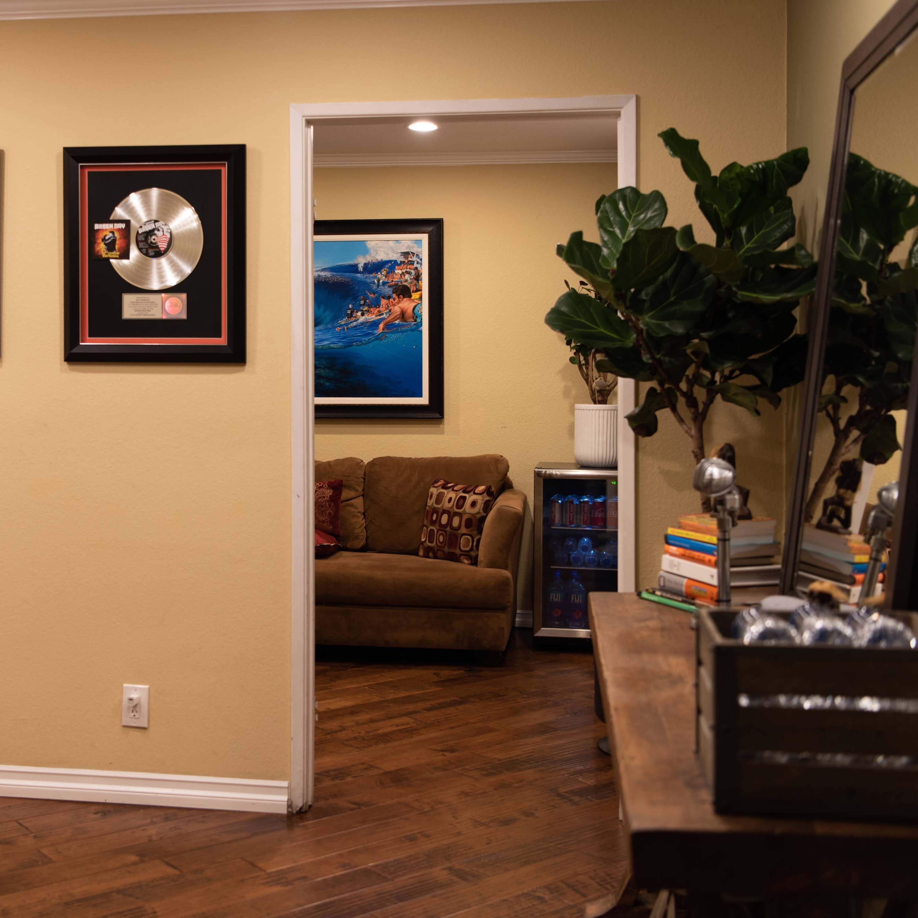 A cozy home office featuring gold and platinum record plaques on the wall, a wooden desk with a tablet stand, and a doorway leading to a living area with a couch and a plant nearby.