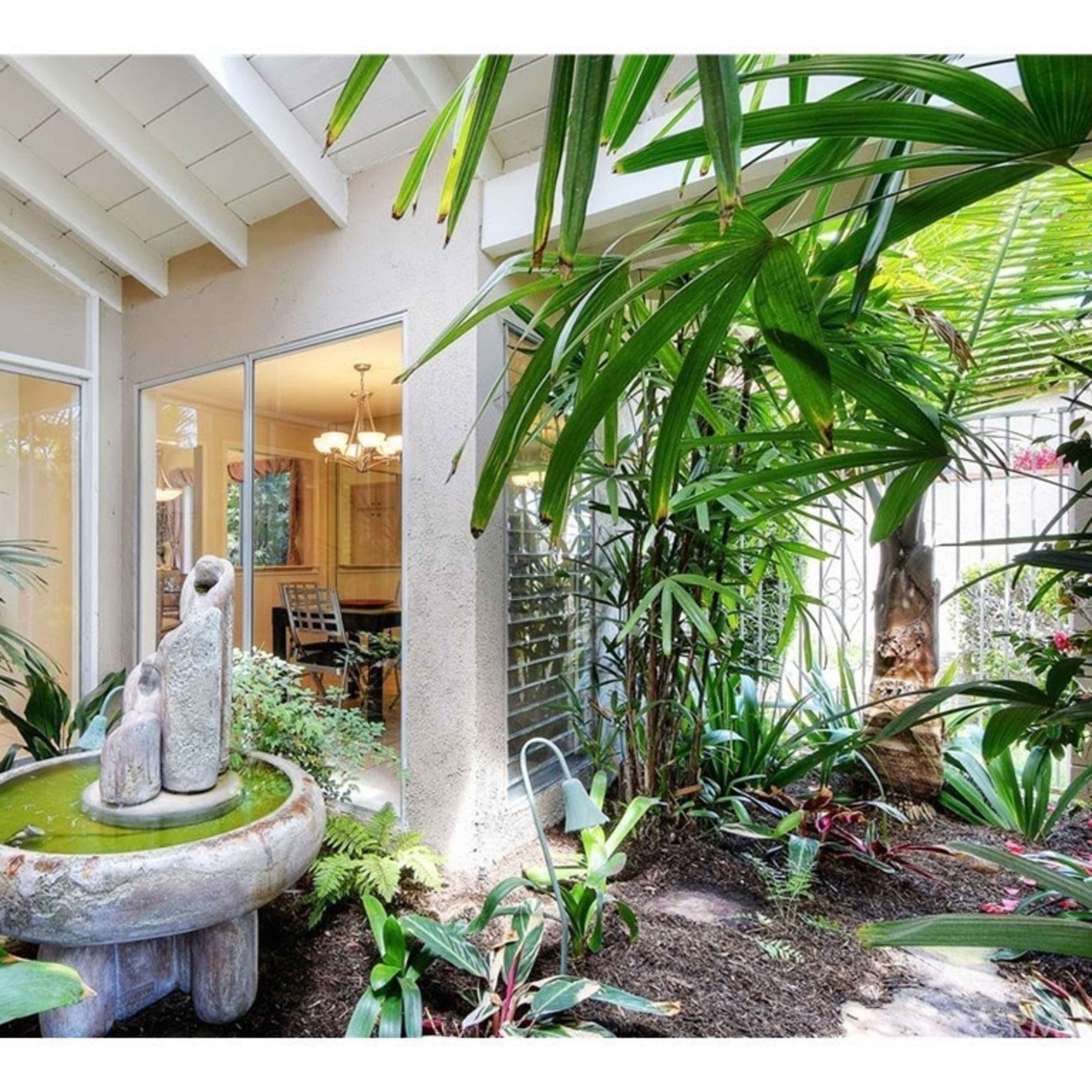 A stone fountain is centered in a lush garden surrounded by tropical plants, with a view of an adjacent room through large glass windows.