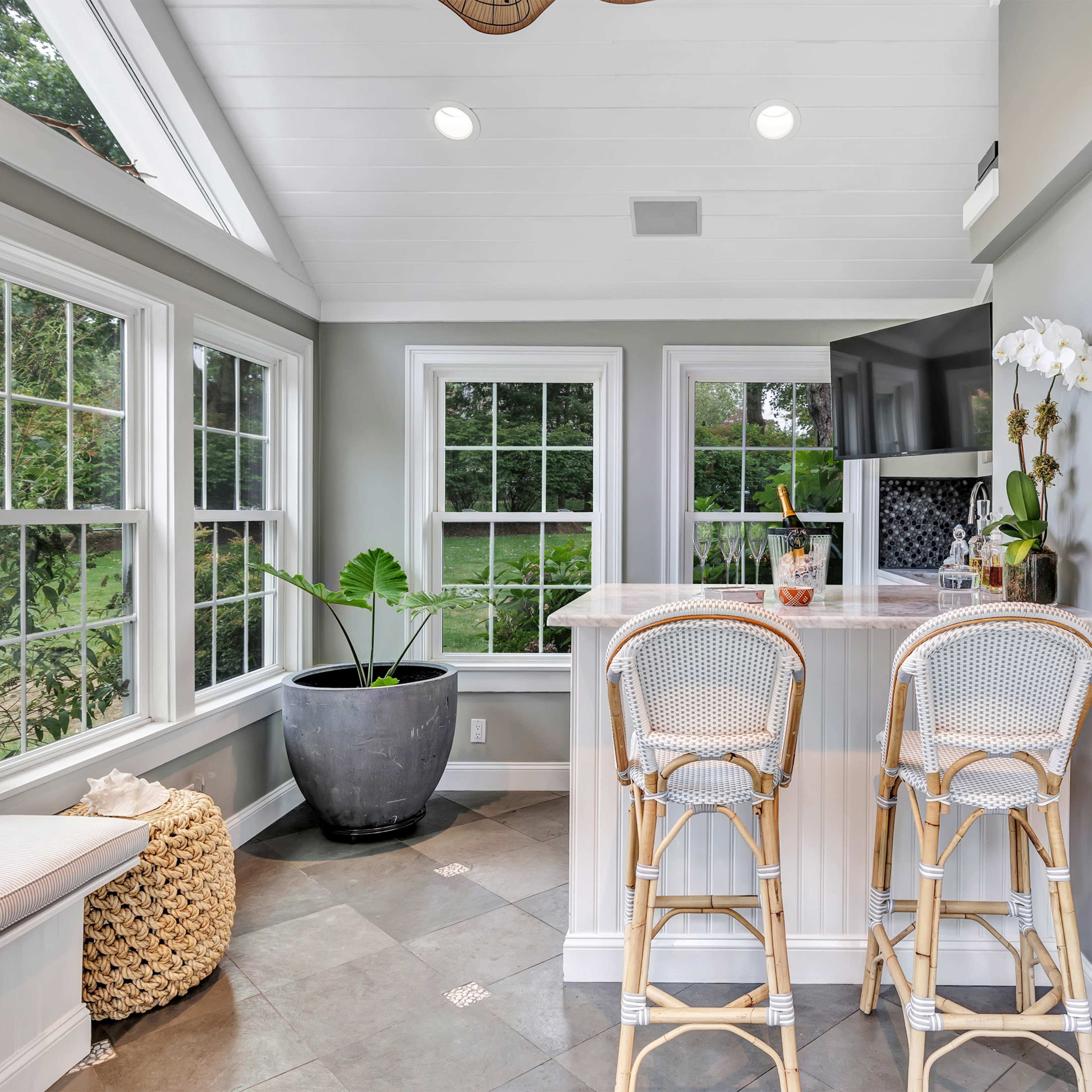 A bright and modern kitchen nook with two rattan bar stools facing a small counter, surrounded by large windows and a decorative potted plant.