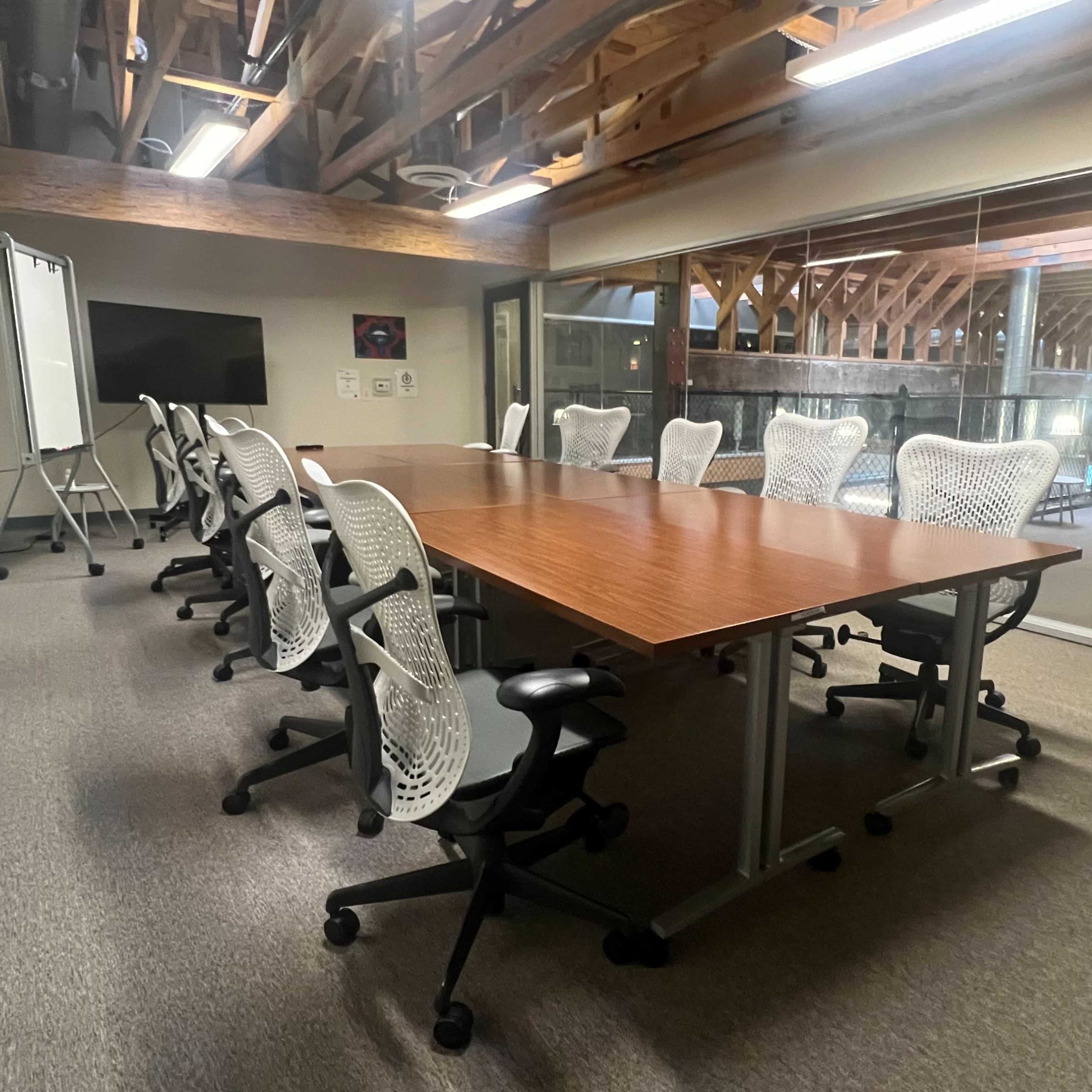 A spacious conference room features a large wooden table surrounded by ergonomic chairs, with large windows revealing a view of a wooden-beamed structure.