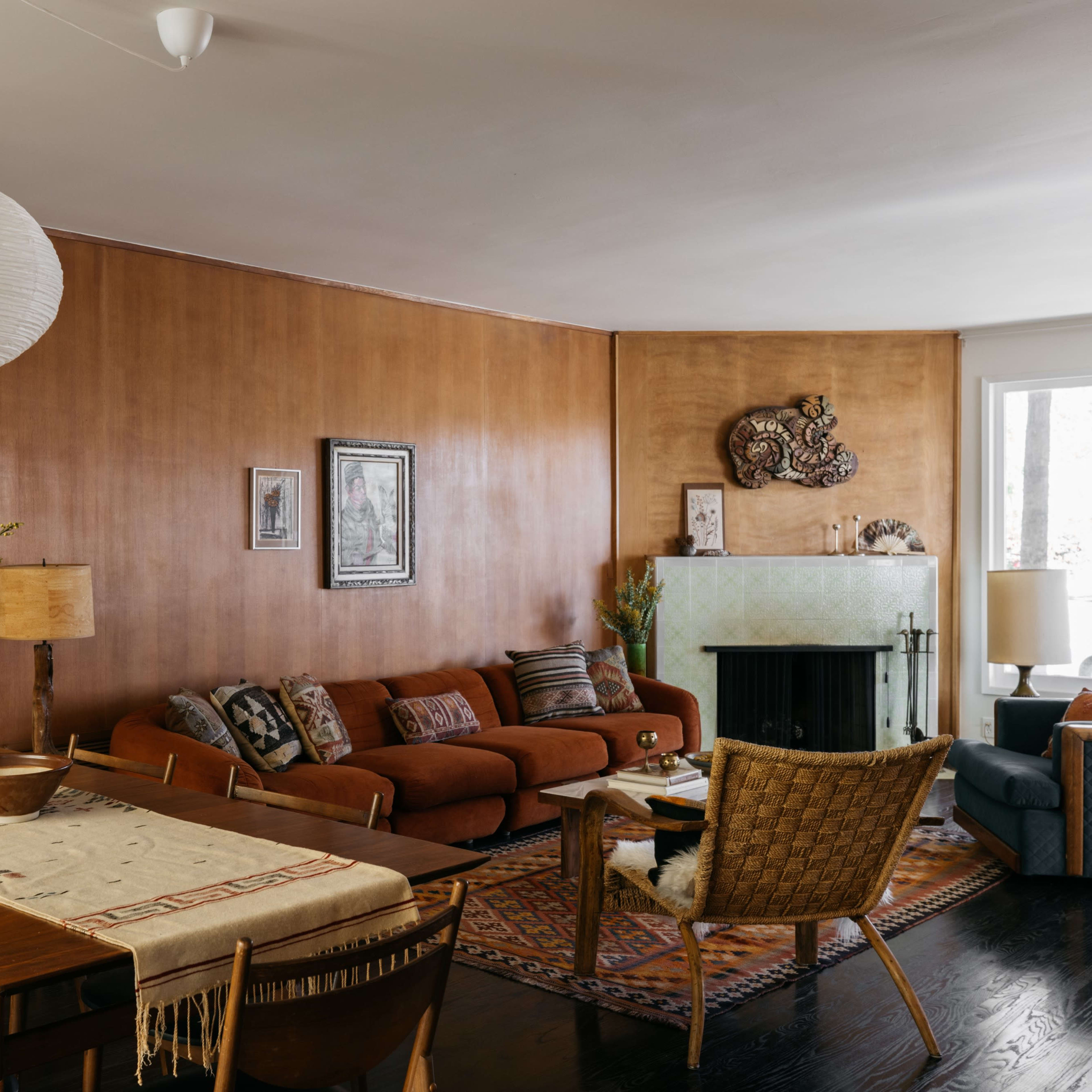A cozy living room with a brown wall, a wooden dining table, a sofa, and a fireplace, all arranged around a patterned rug.