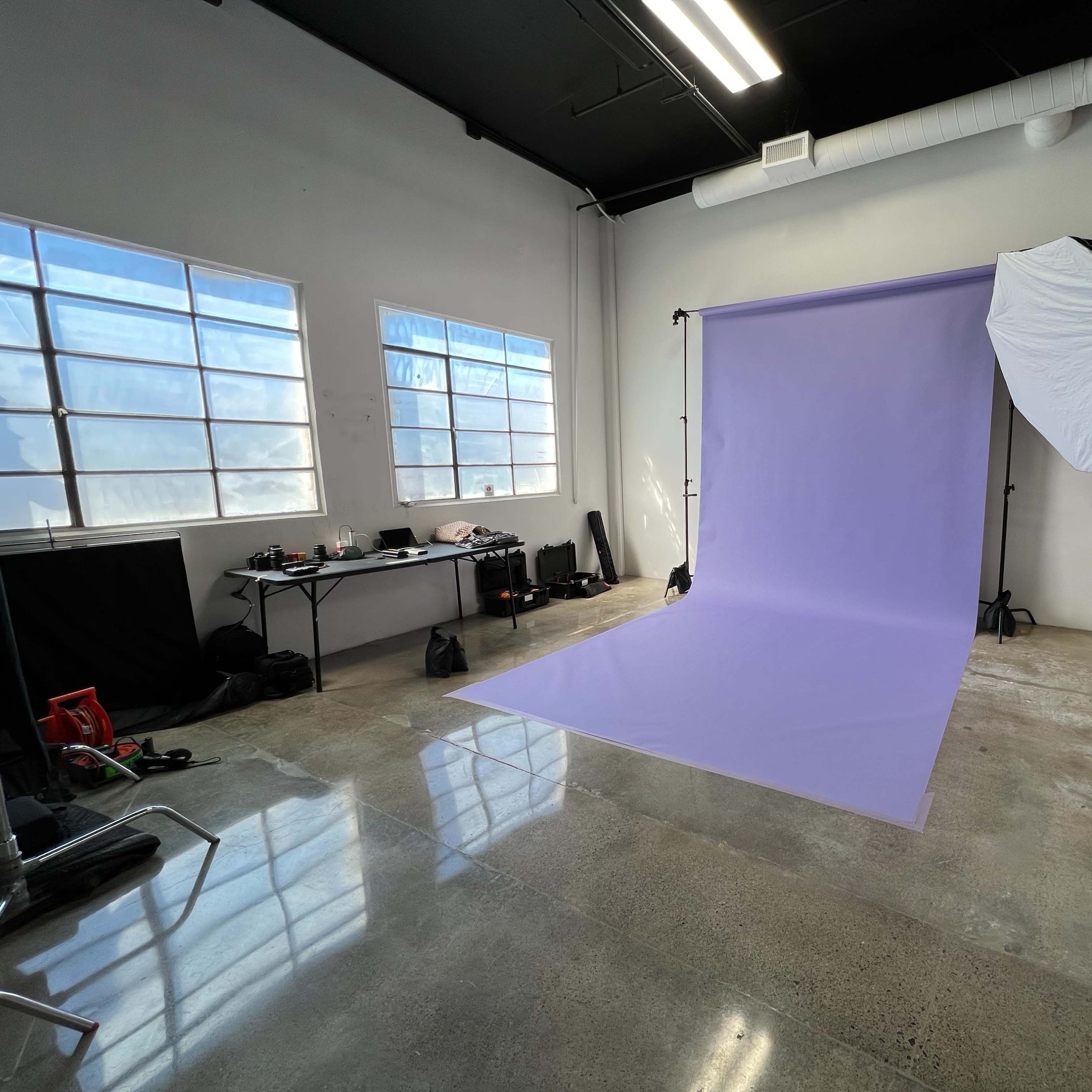 A photography studio features a lavender backdrop on a polished concrete floor, with large windows illuminating the space and various equipment visible in the background.